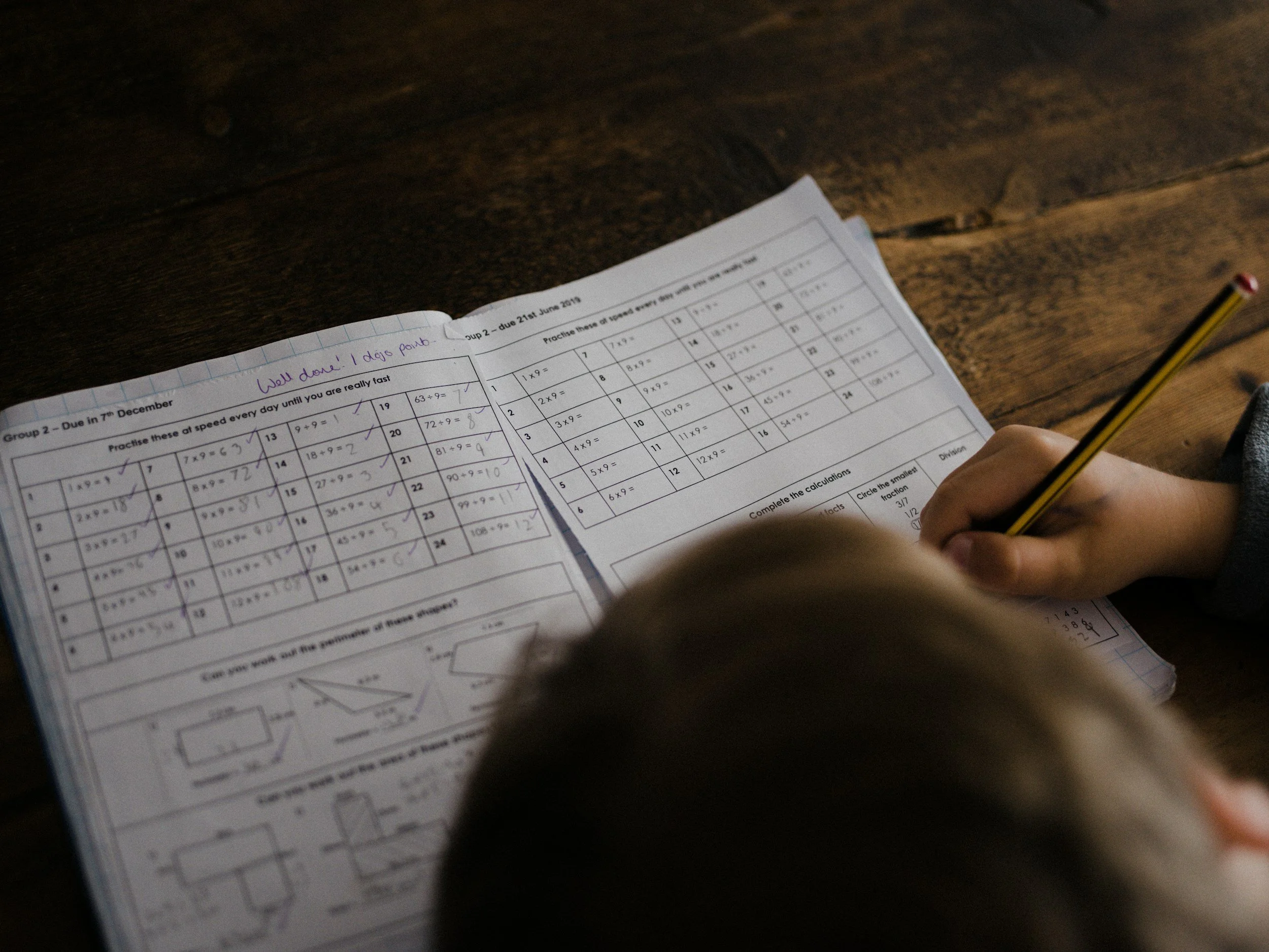 Child completing math worksheets at a wooden table, with a pencil in hand and a smiley face and date written on the paper.