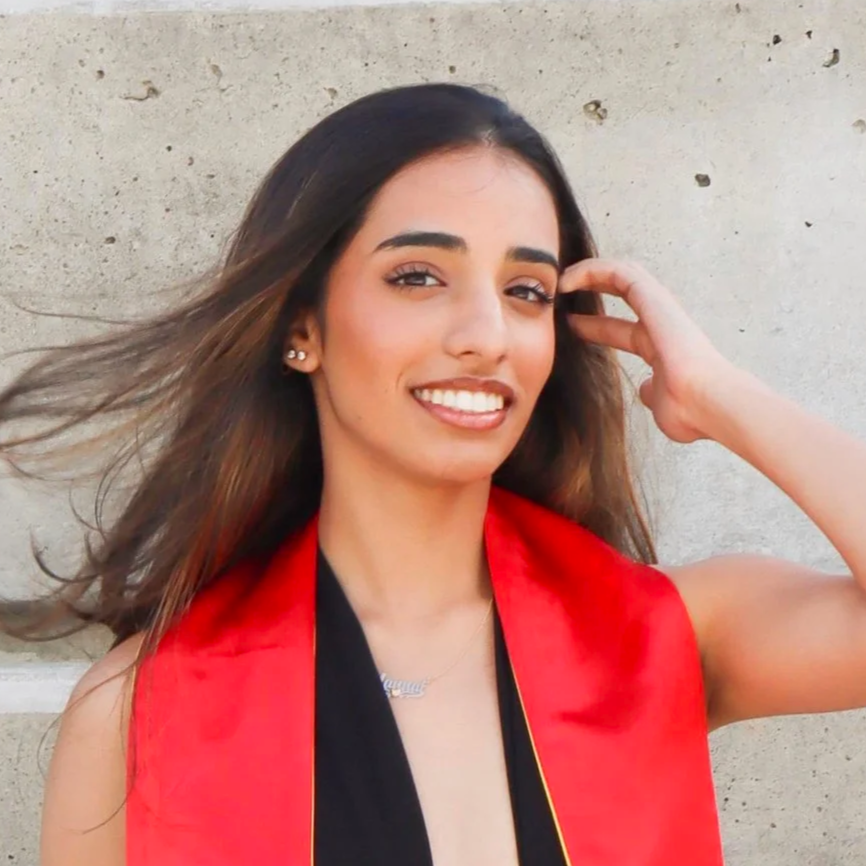 A young woman with long hair smiling, wearing a red and black stole, standing against a concrete wall.