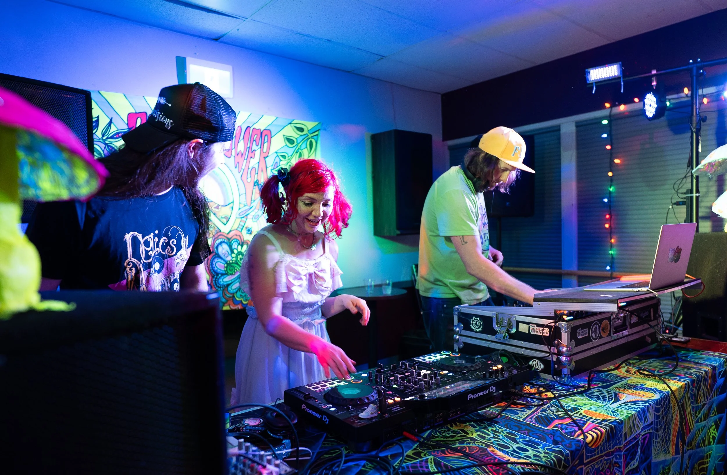 Three people performing as DJs at a colorful event with neon lights. One woman and two men are behind DJ equipment, with the woman in the middle wearing a white dress and the man on the right wearing a yellow cap. The background has a vibrant, psyche