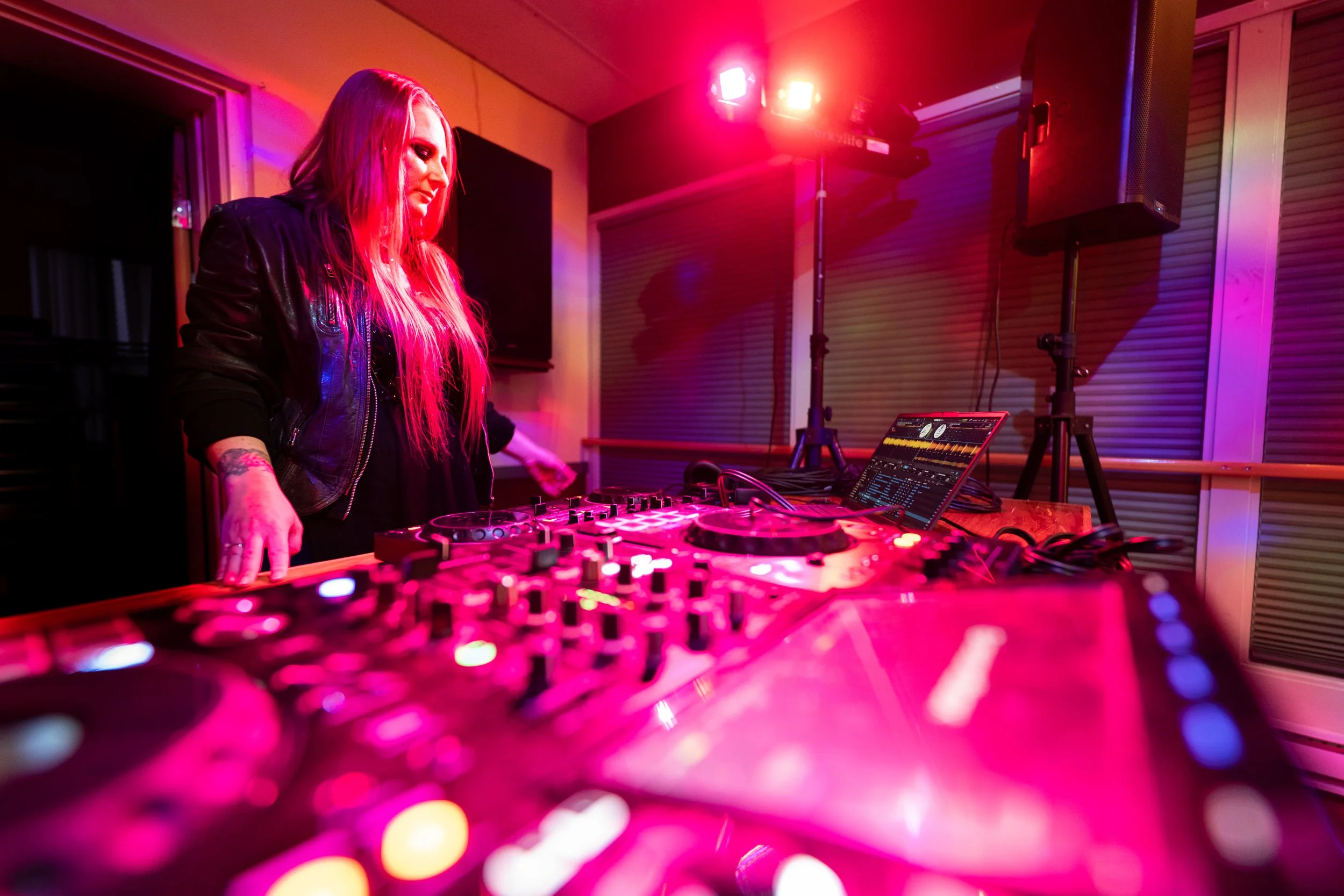 A DJ with long blonde hair wearing a black leather jacket performing at a party with colorful pink and purple lighting. She is using DJ equipment with a laptop and a mixer in front of her.