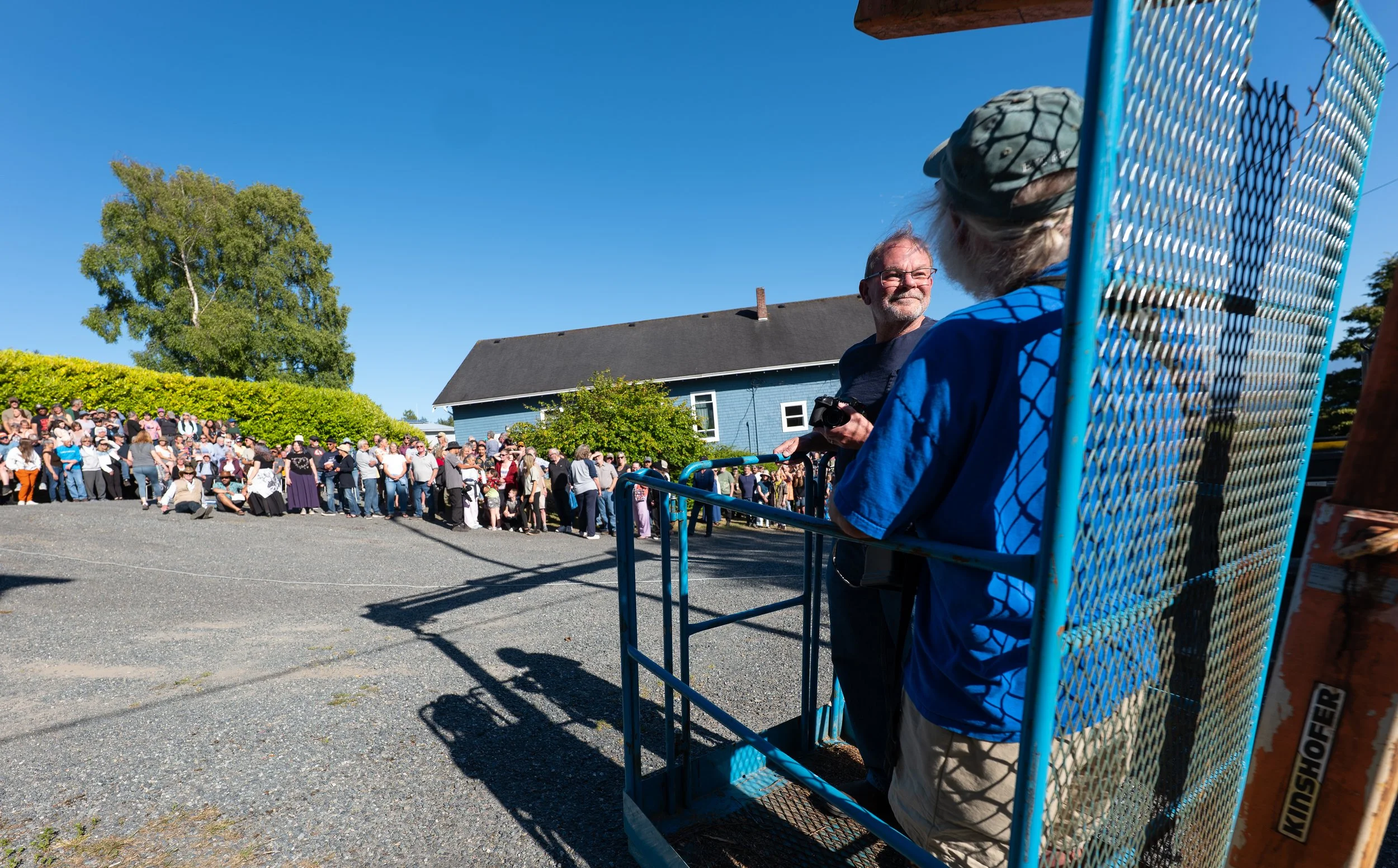 A large crowd of people outside on a sunny day, some standing and some kneeling, with a man and a woman in the foreground near a blue gate. The man is holding a camera and the woman is sitting on the gate, facing away from the camera. There is a hous