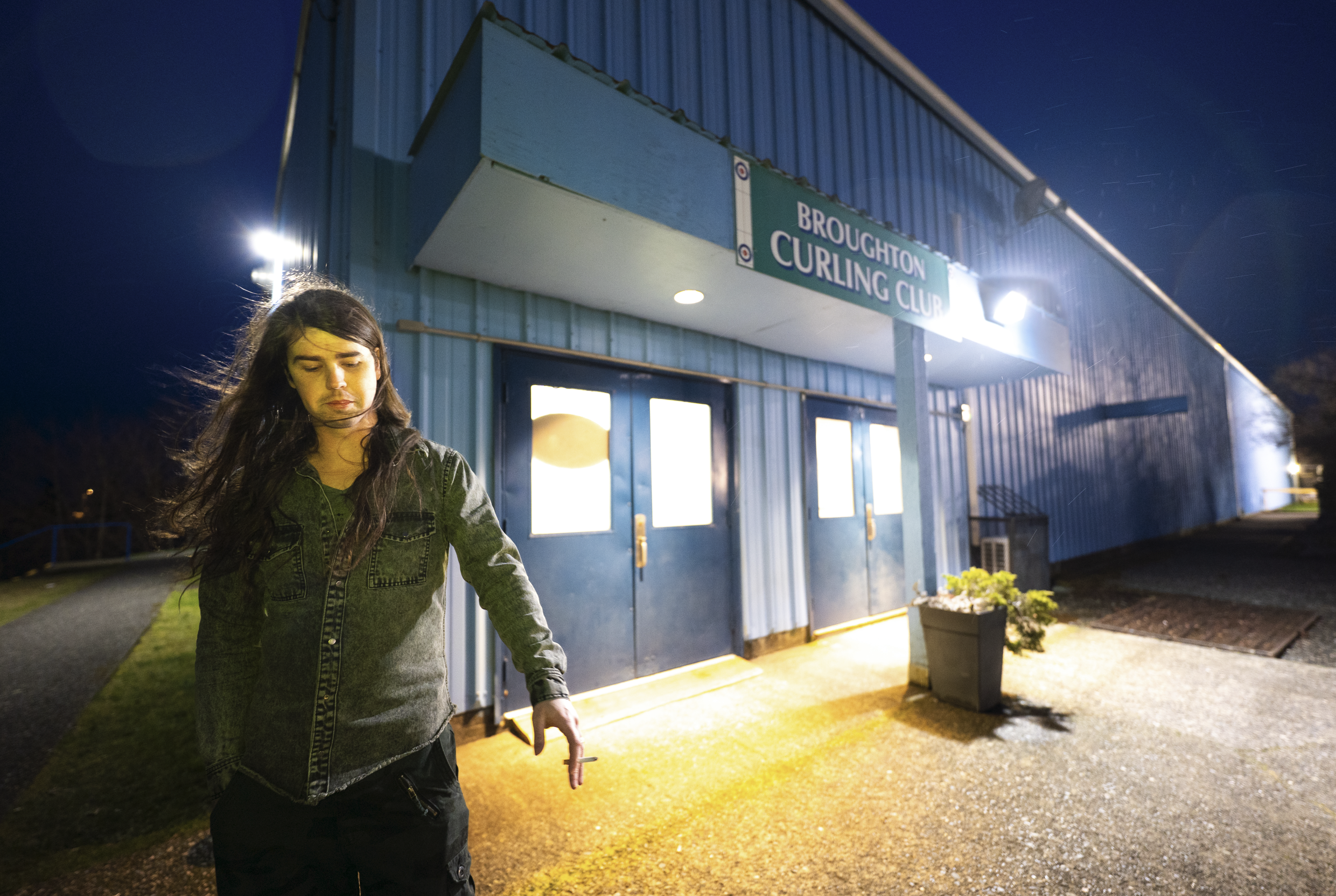 A  with long dark hair, wearing a denim jacket, is walking outside at night in front of a blue building with a sign that reads 'Brough ton Curling Club.' The building is lit by exterior lights, and the woman appears to be focused on what she's doing.