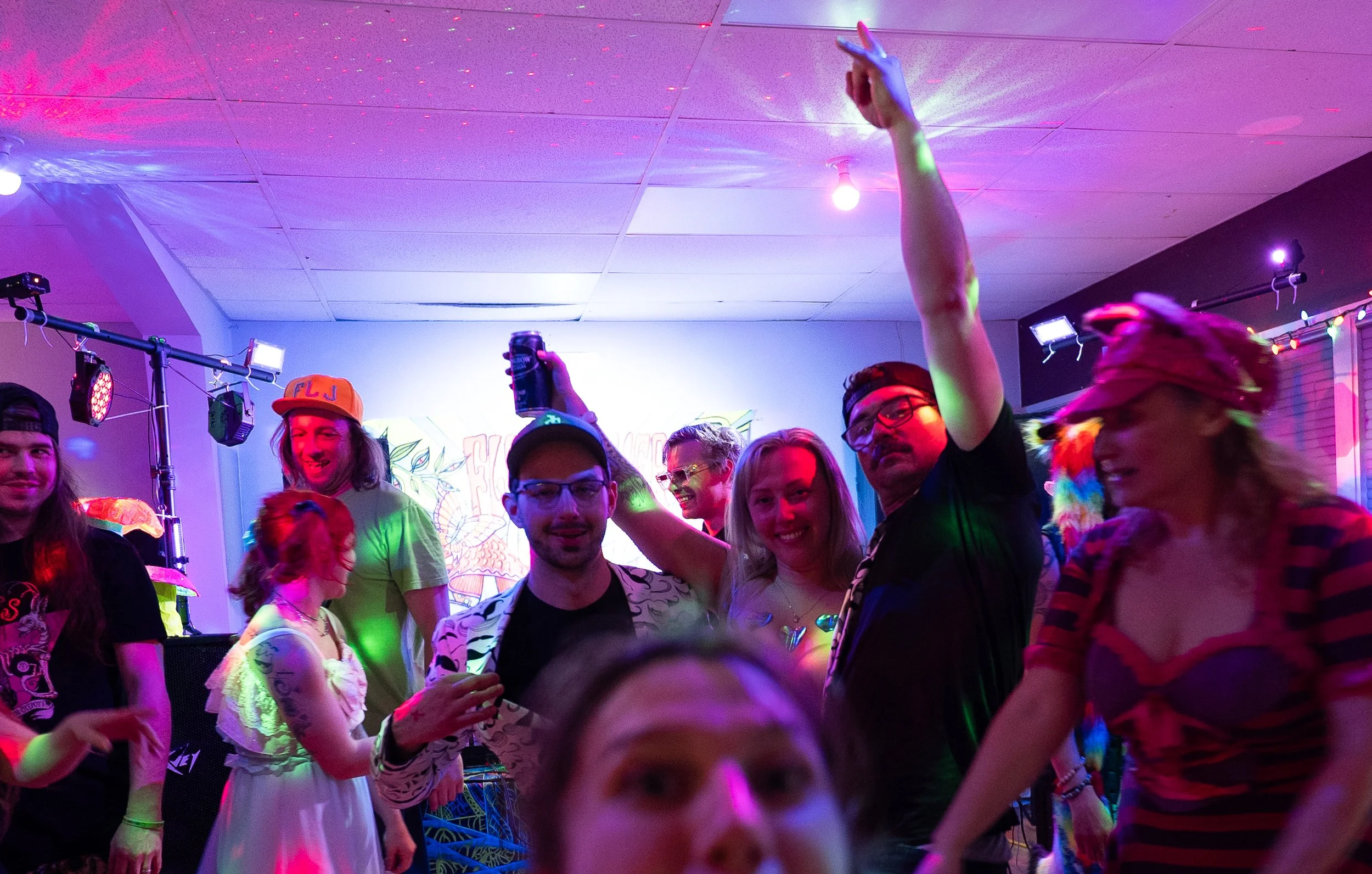 People dancing and enjoying a party under colorful lighting, with some holding drinks, in a lively indoor setting.