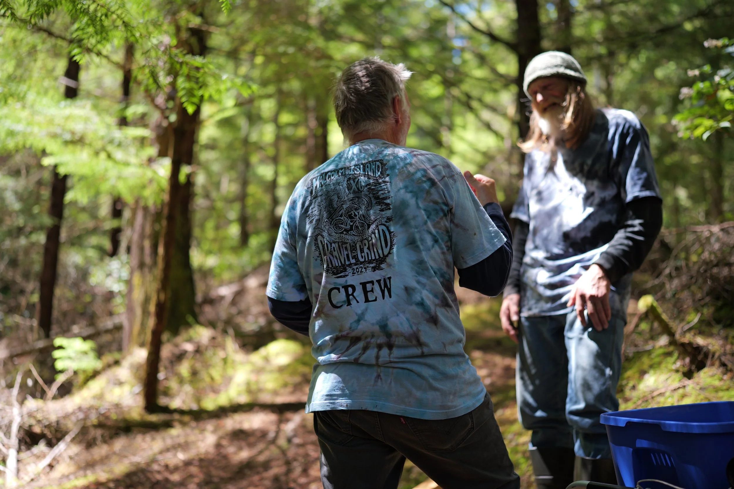 Two people having a conversation in a forested area, one wearing a tie-dye shirt with a printed design and the other in a hat and tie-dye shirt. The back of the person in the tie-dye shirt is visible, and the person's shirt has the words 'Welcome to 