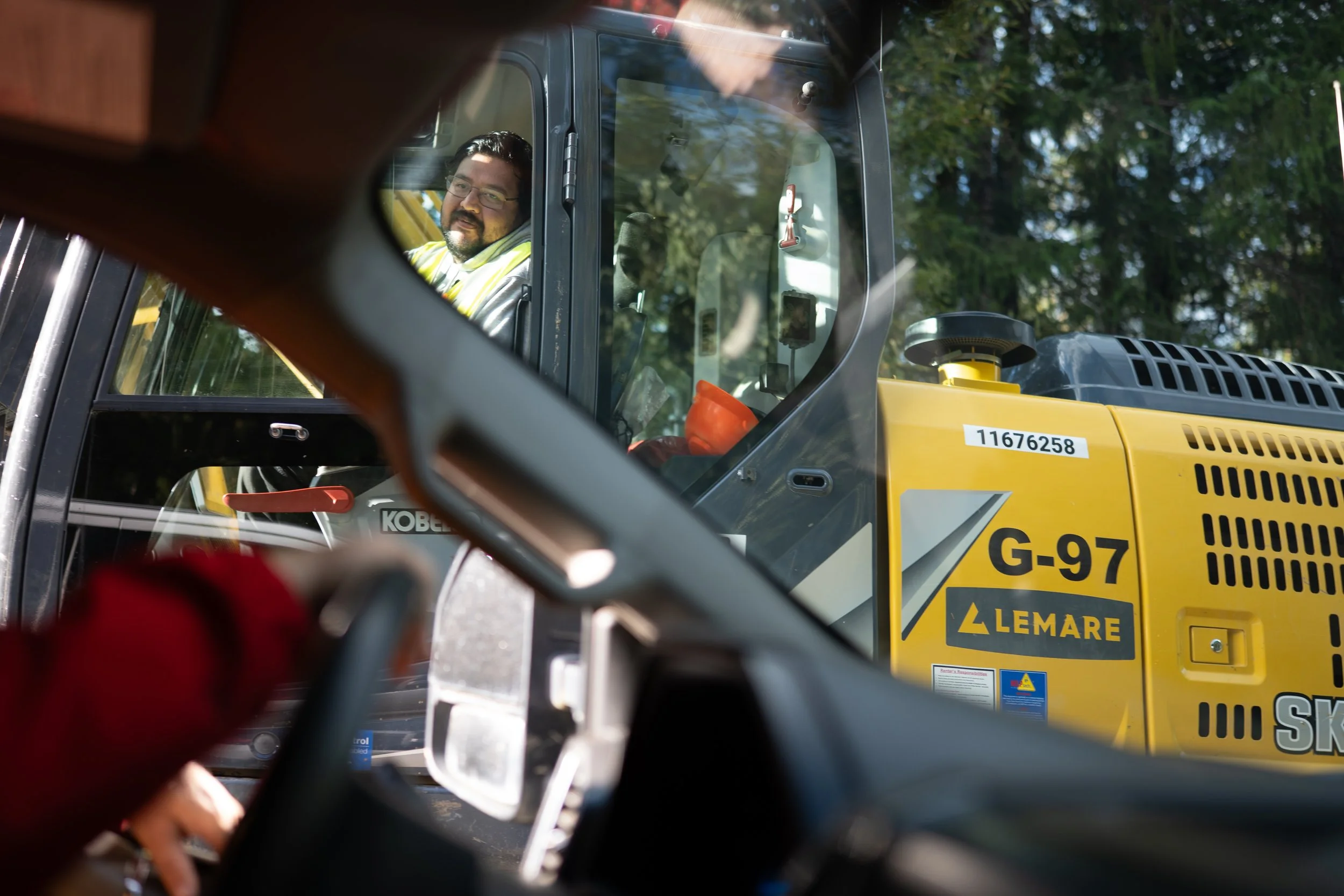 A man with glasses and a beard is sitting inside a construction vehicle, looking out through the window, with trees in the background.