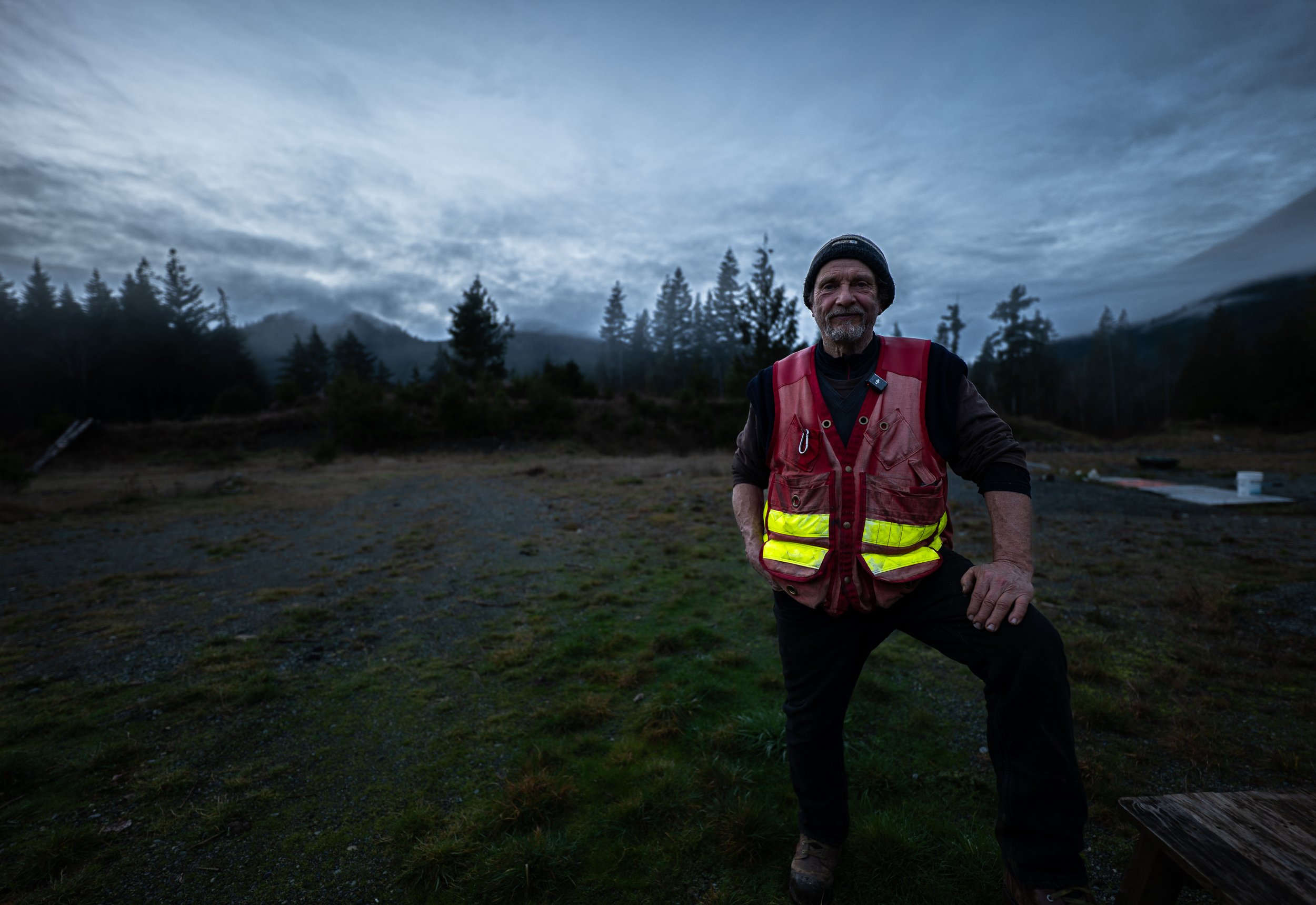 An man with a beard wearing a black beanie and a red safety vest standing outdoors in a forested area under a cloudy sky.