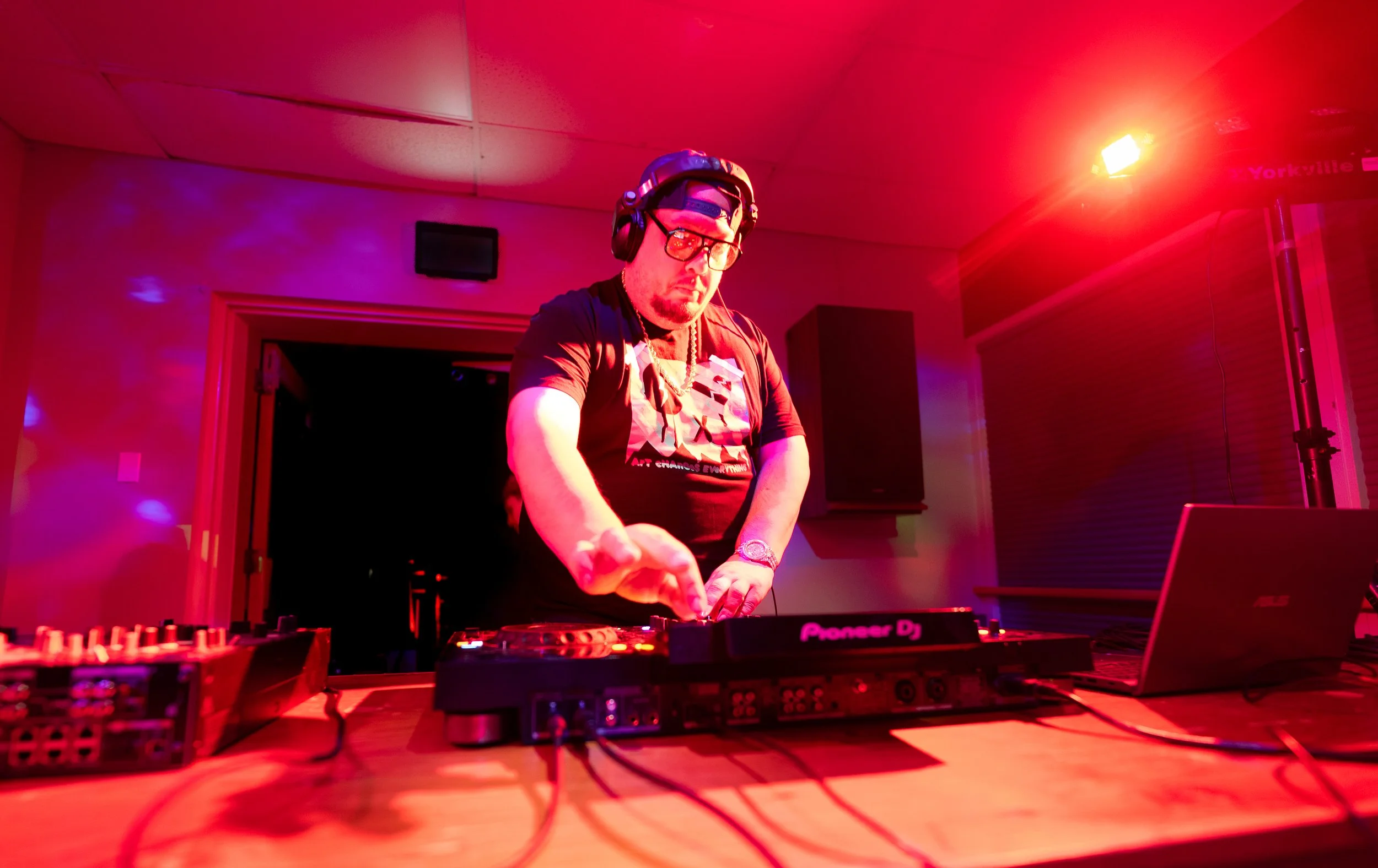 DJ wearing headphones, glasses, and a chain, performing with music equipment in a dimly lit room with red and purple lighting.