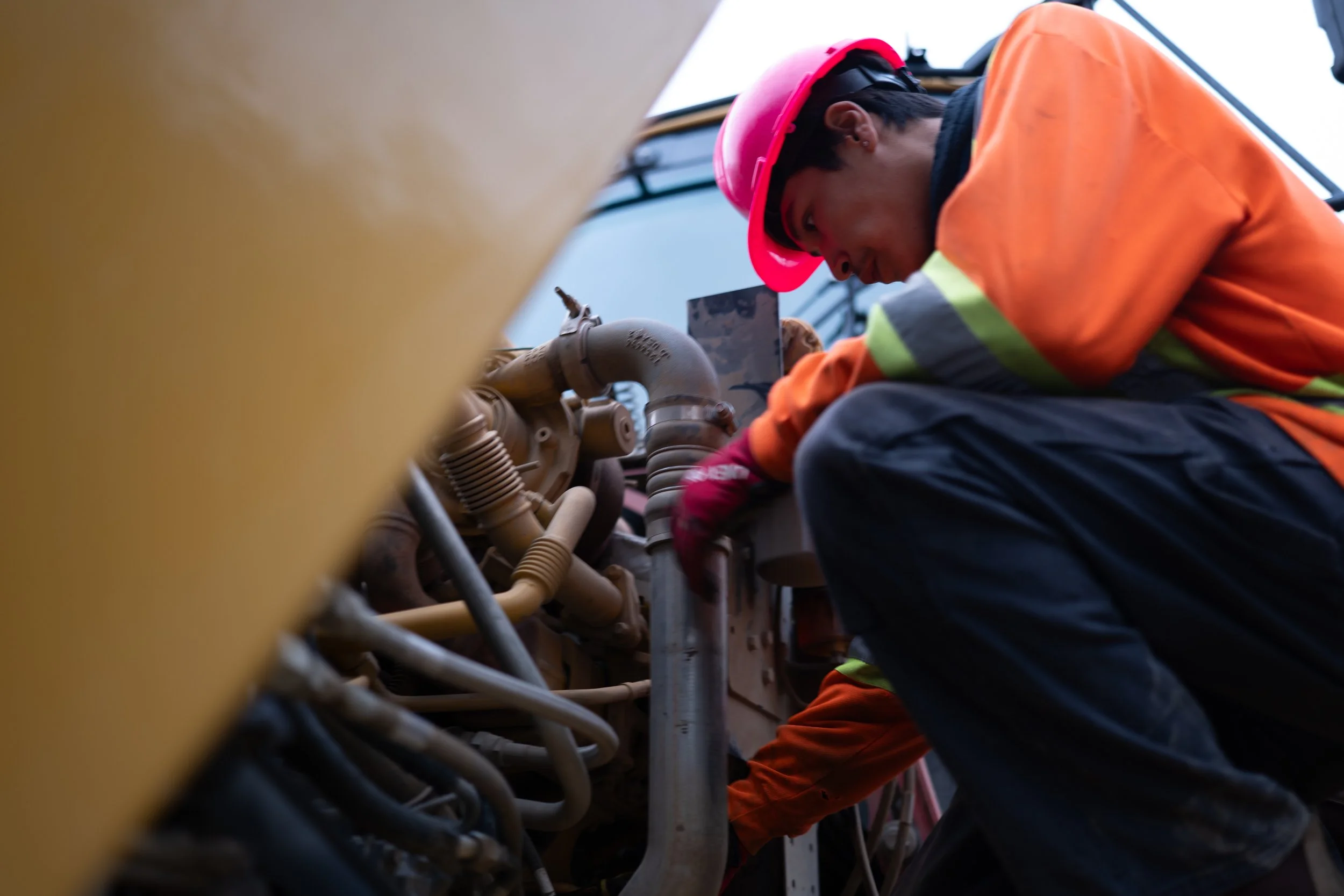 Worker in safety gear, including a red helmet and orange jacket, working on machinery with pipes and valves during daytime.
