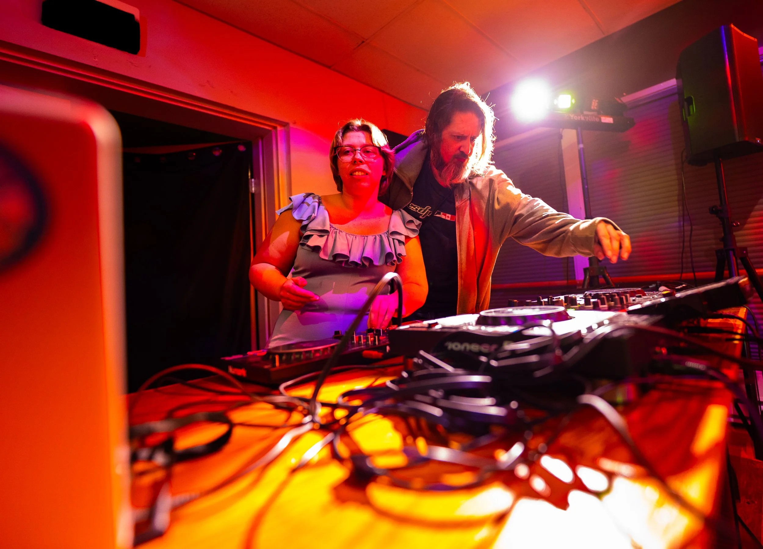 Two DJs at a party with colorful lighting, surrounded by DJ equipment and cables.