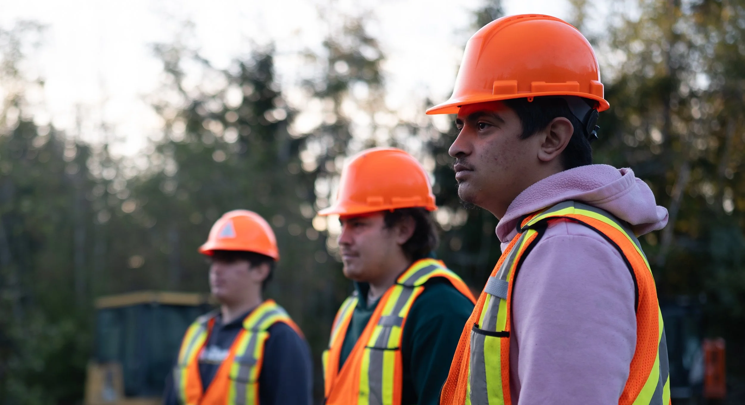 Group of young men wearing orange safety helmets and reflective vests standing outdoors in a line, with trees in the background.