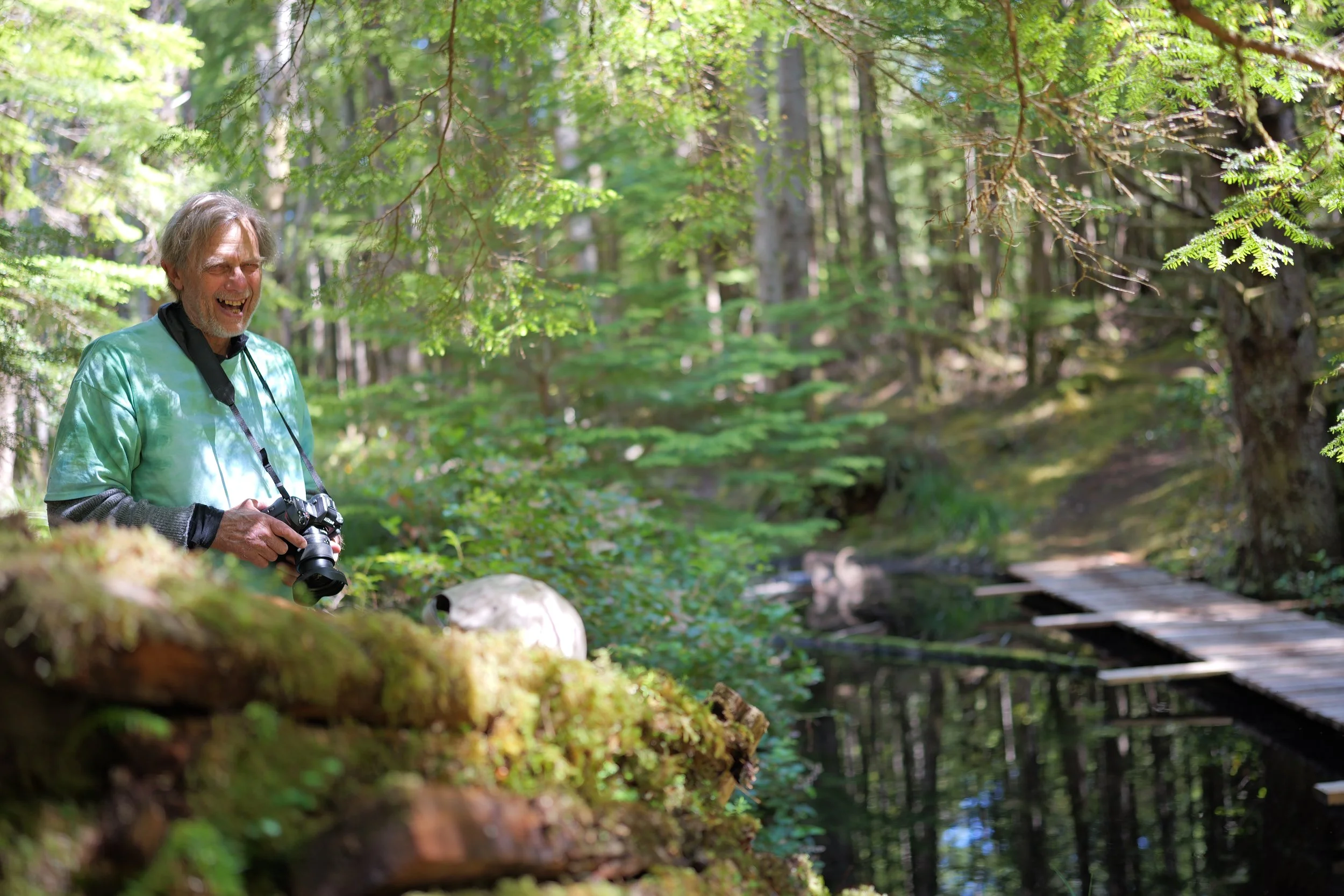 A man  wearing a green shirt and black gloves, holding a camera, standing in a lush green forest near a small wooden bridge over a creek, smiling and enjoying the outdoor scenery.