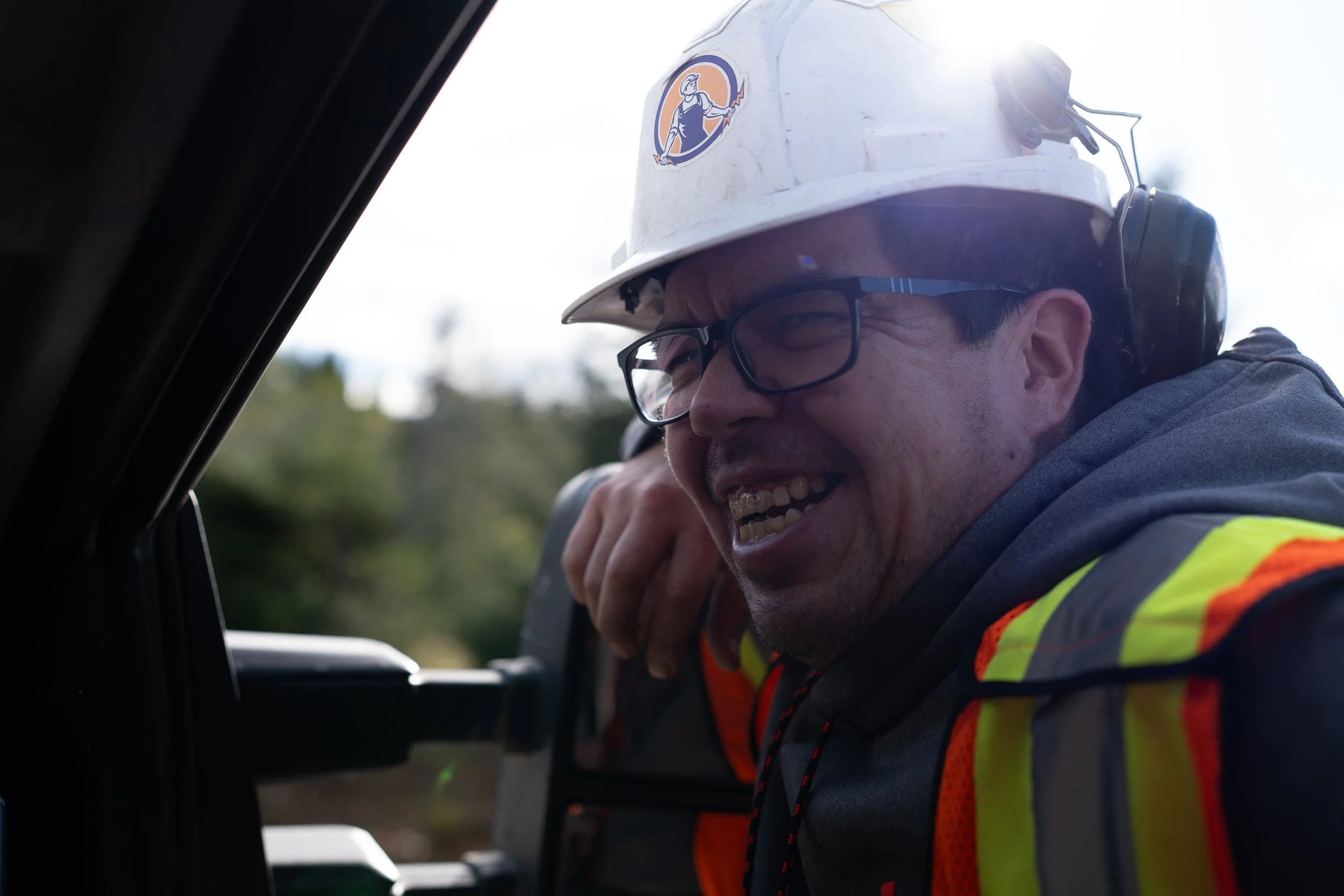 A construction worker wearing a white hard hat with a logo, glasses, and a high-visibility safety vest is smiling and leaning into the window of a vehicle.