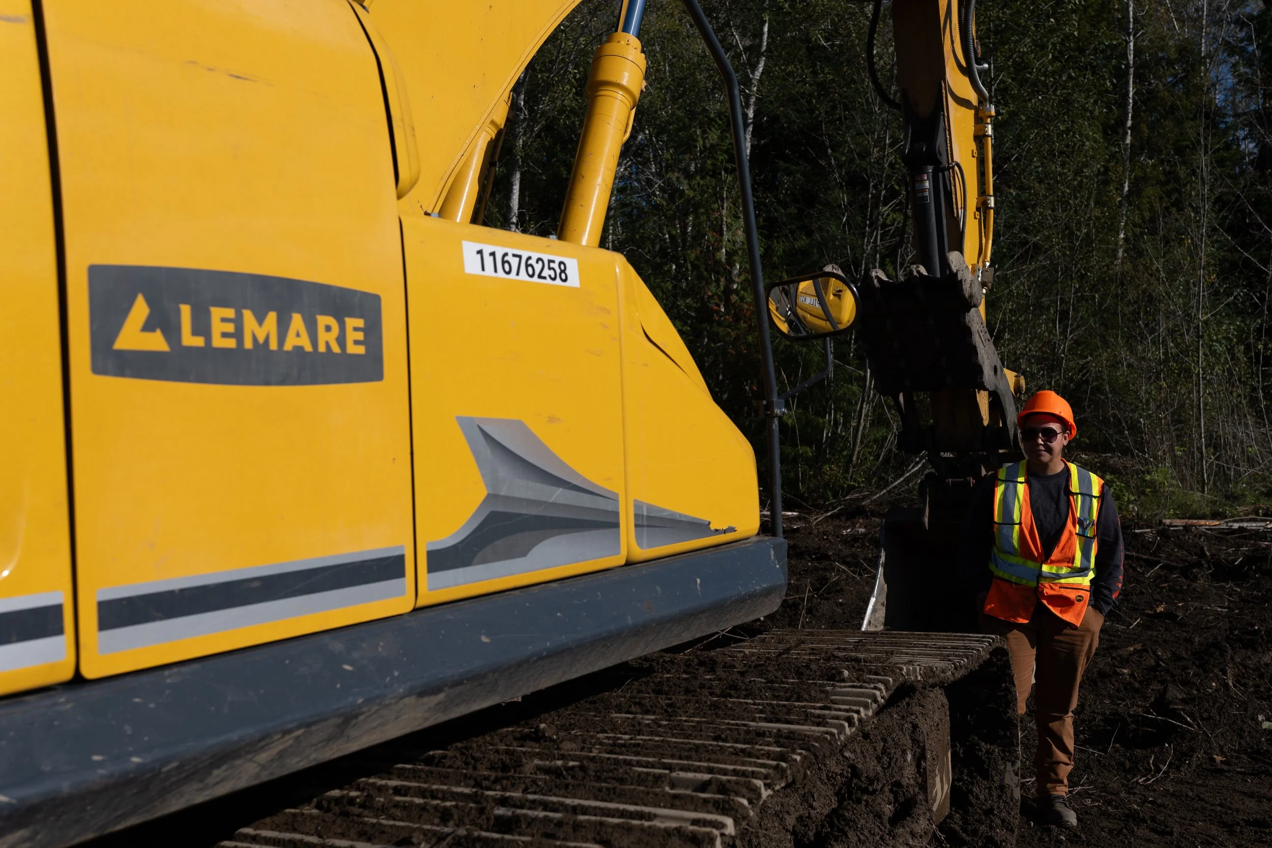 An HEO student wearing a safety vest and orange helmet walking beside a large yellow excavator in a forested area with trees and sunlight.