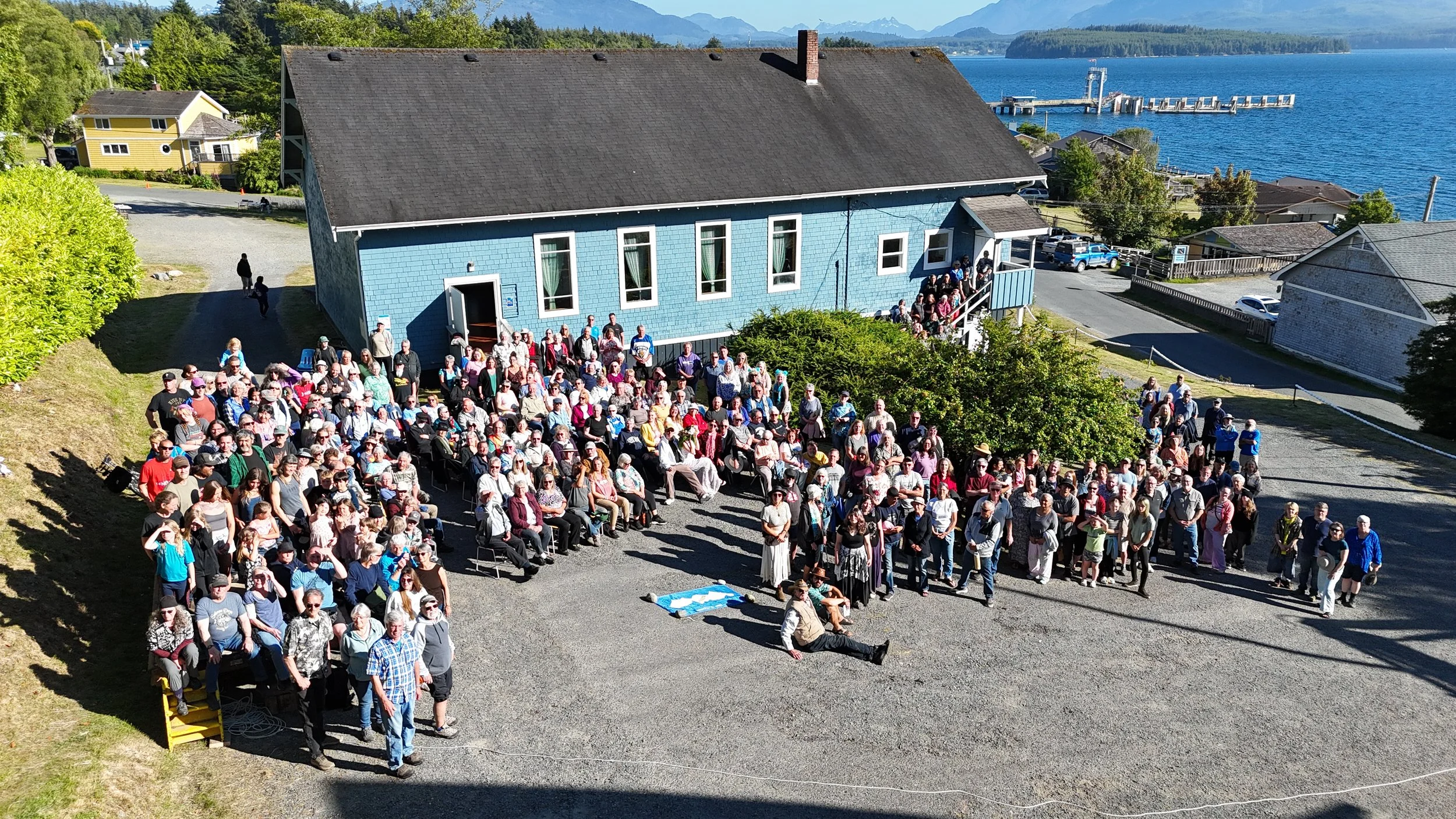 A large outdoor gathering with many people seated and standing in front of a blue building by a body of water, with trees and mountains in the background on a sunny day.