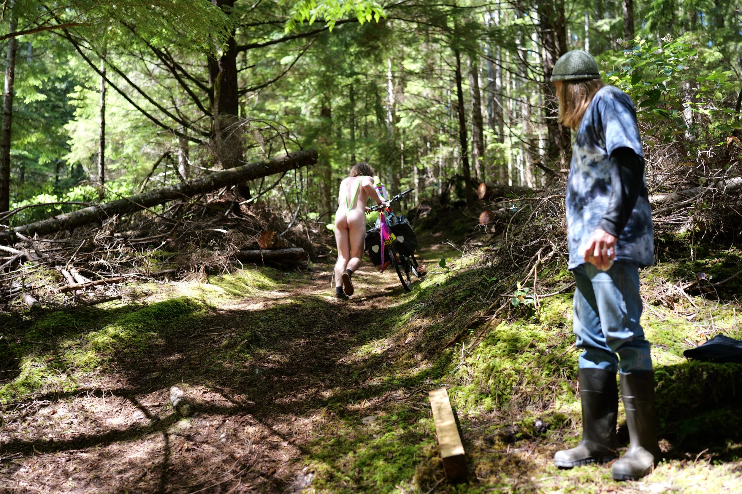 A cyclist, partly dressed, pushing a bicycle along a dirt path surrounded by trees and greenery. Another person, wearing a hat and rain boots, stands nearby.