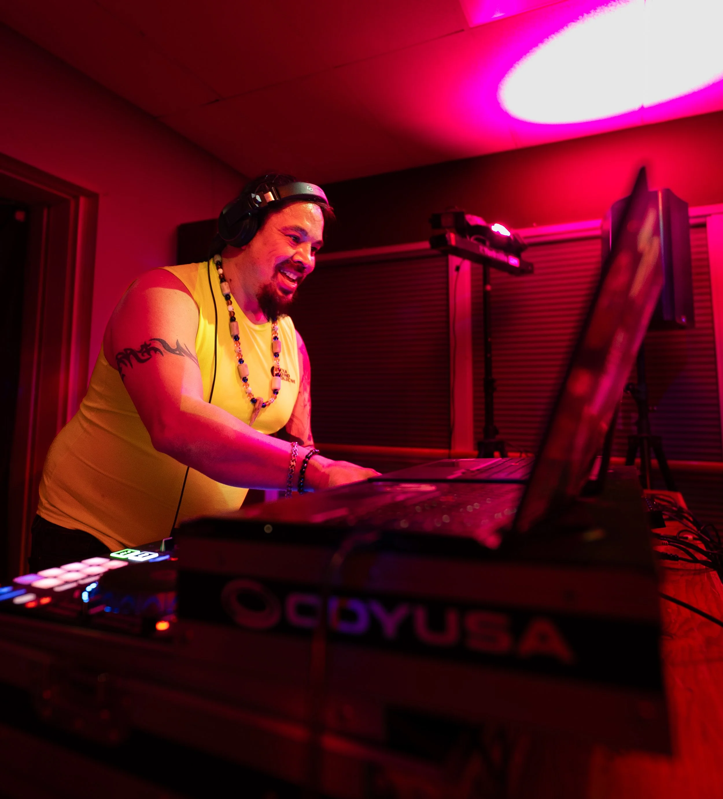 A DJ wearing headphones and a yellow tank top is smiling while DJing, with DJ equipment and pink lighting in the background.