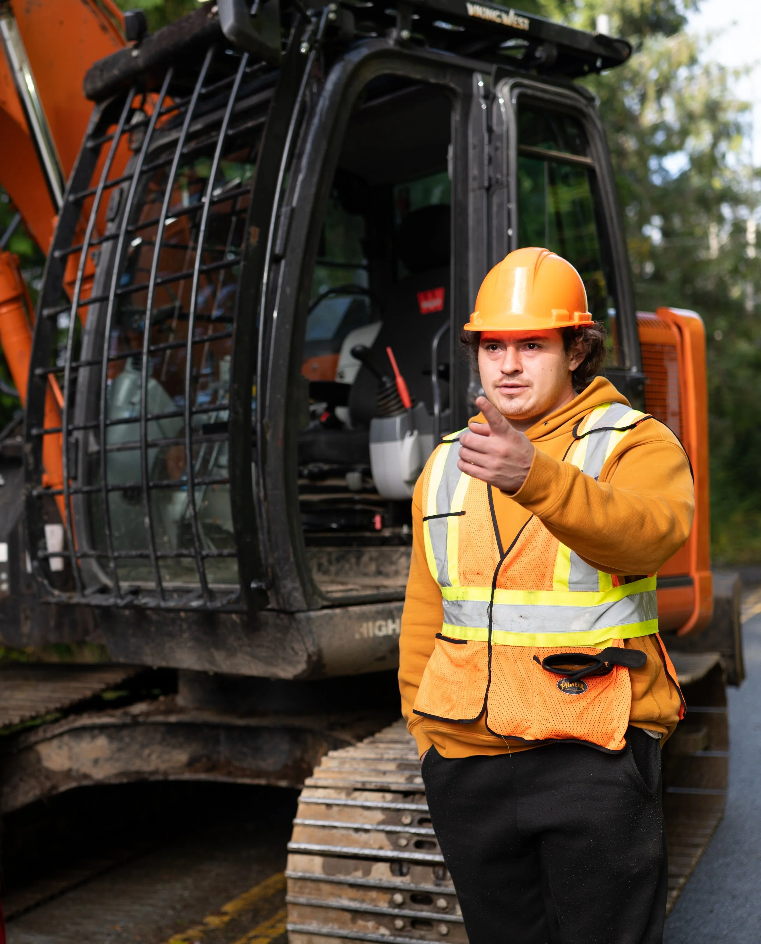 A construction worker wearing an orange hard hat, yellow safety vest, and black pants standing in front of construction equipment, pointing and looking to the side.