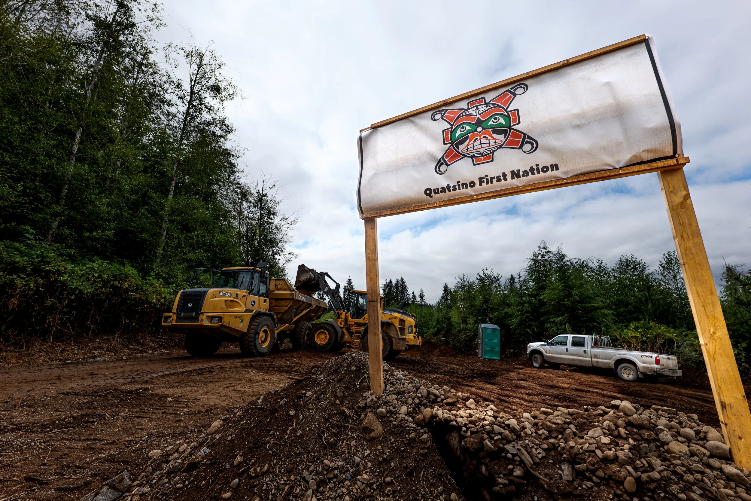 Construction site with a large sign that reads 'Quatsino First Nation,' showing a colorful Indigenous-inspired design. Heavy machinery and vehicles parked on uneven dirt ground, surrounded by green trees under cloudy sky.