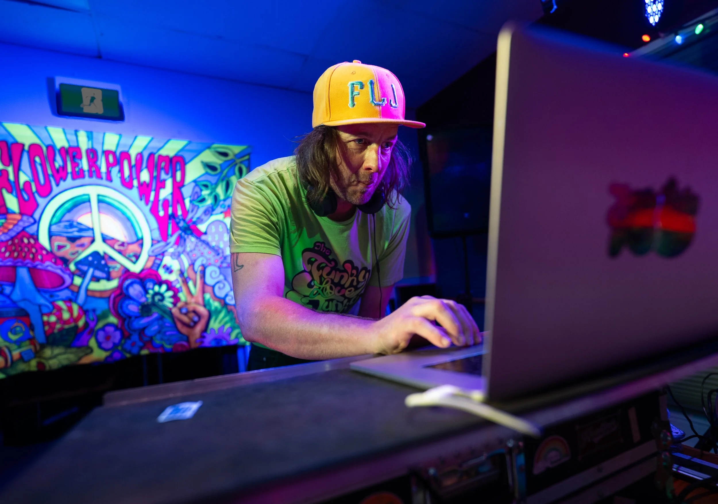 A man with long hair wearing a yellow cap and headphones focuses on a DJ setup, with colorful psychedelic artwork and a peace sign in the background.