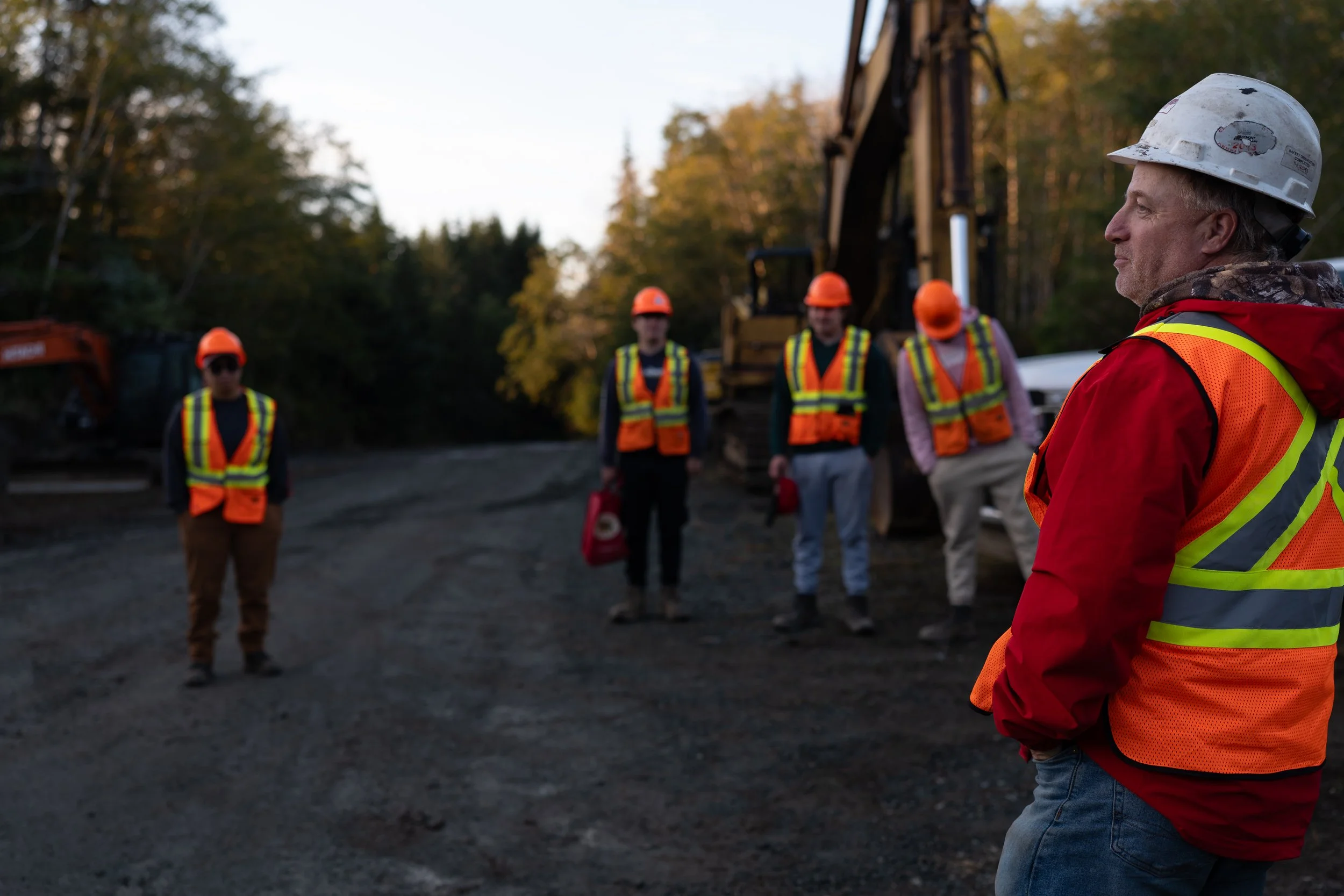 A group of construction workers standing outdoors wearing safety helmets and reflective vests, with one worker in the foreground facing right and the others in the background.