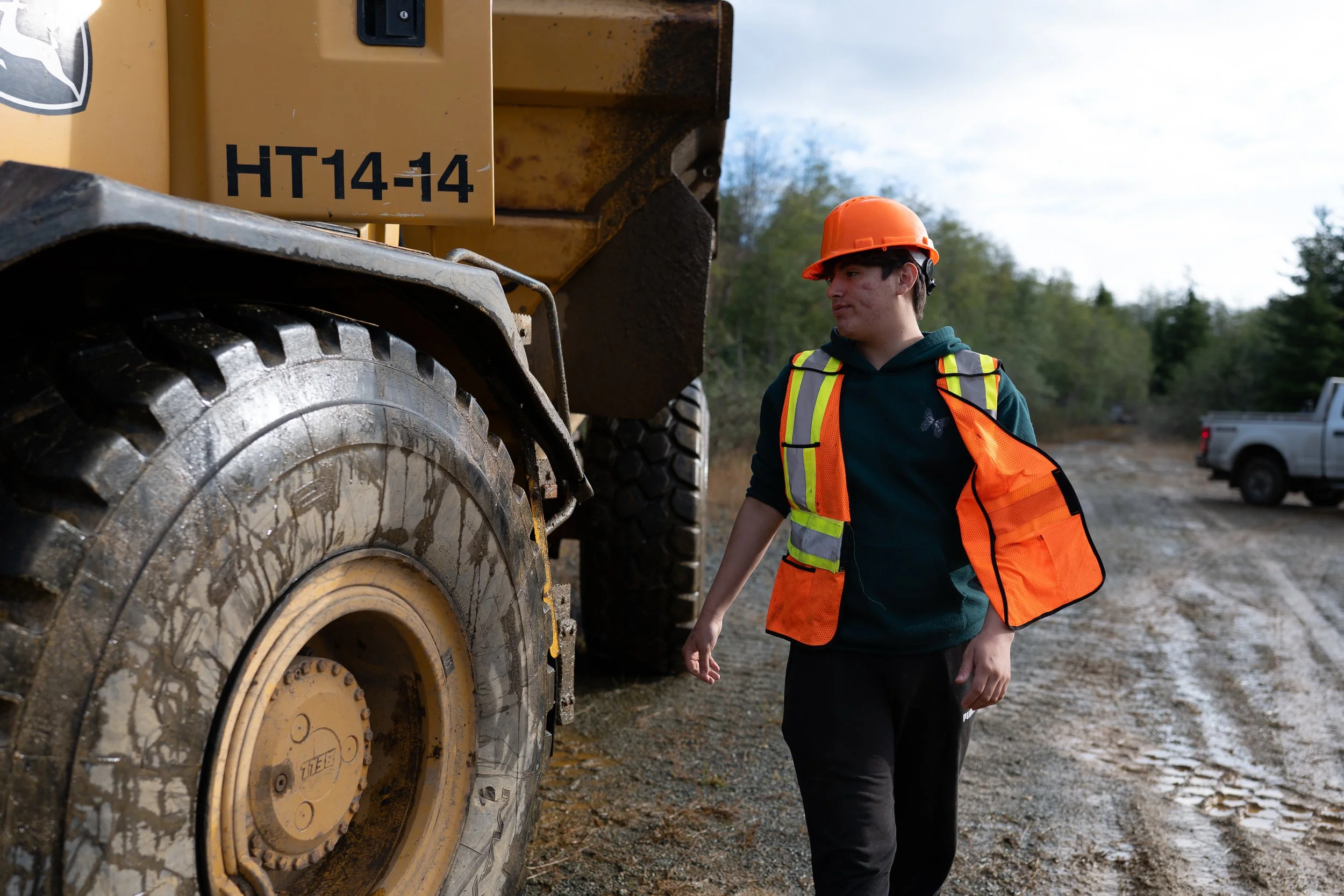 A construction worker wearing an orange hard hat and a neon safety vest looks at a large, muddy yellow construction vehicle on a dirt road, with trees and a blue sky in the background.