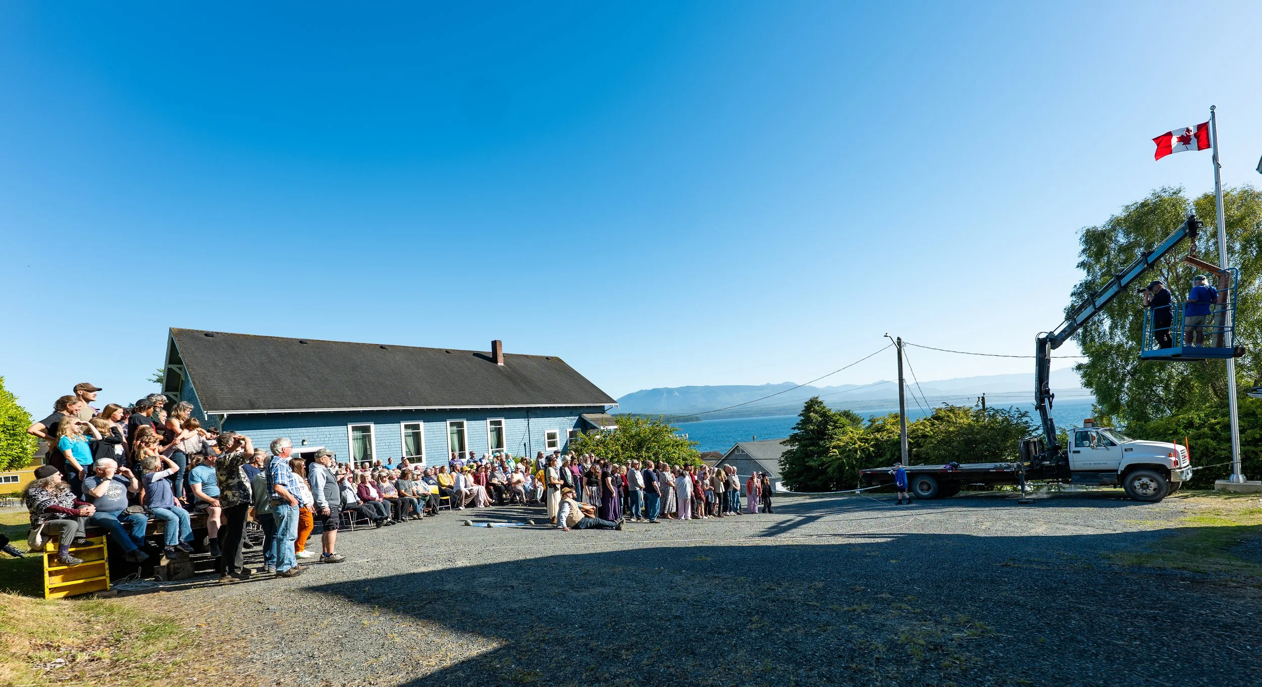A crowd of people gathered outdoors near a blue house with a sloped roof, overlooking a body of water with mountains in the distance. A large flagpole with a Canadian flag is visible, and a cherry picker lift extended high with two individuals workin