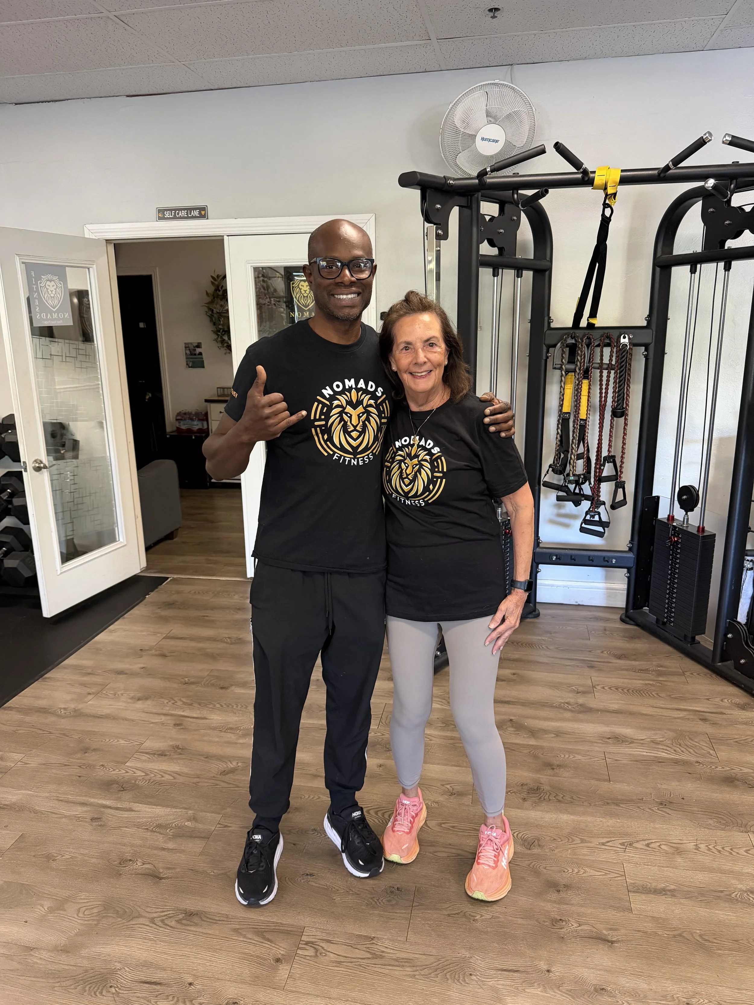A man and woman smiling and posing in a gym, wearing black T-shirts with a lion logo and the words 'Nomads Fitness'. The man is giving a shaka sign with his right hand, and the woman is wearing pink sneakers. There is gym equipment in the background, including a pull-up frame and resistance ropes.