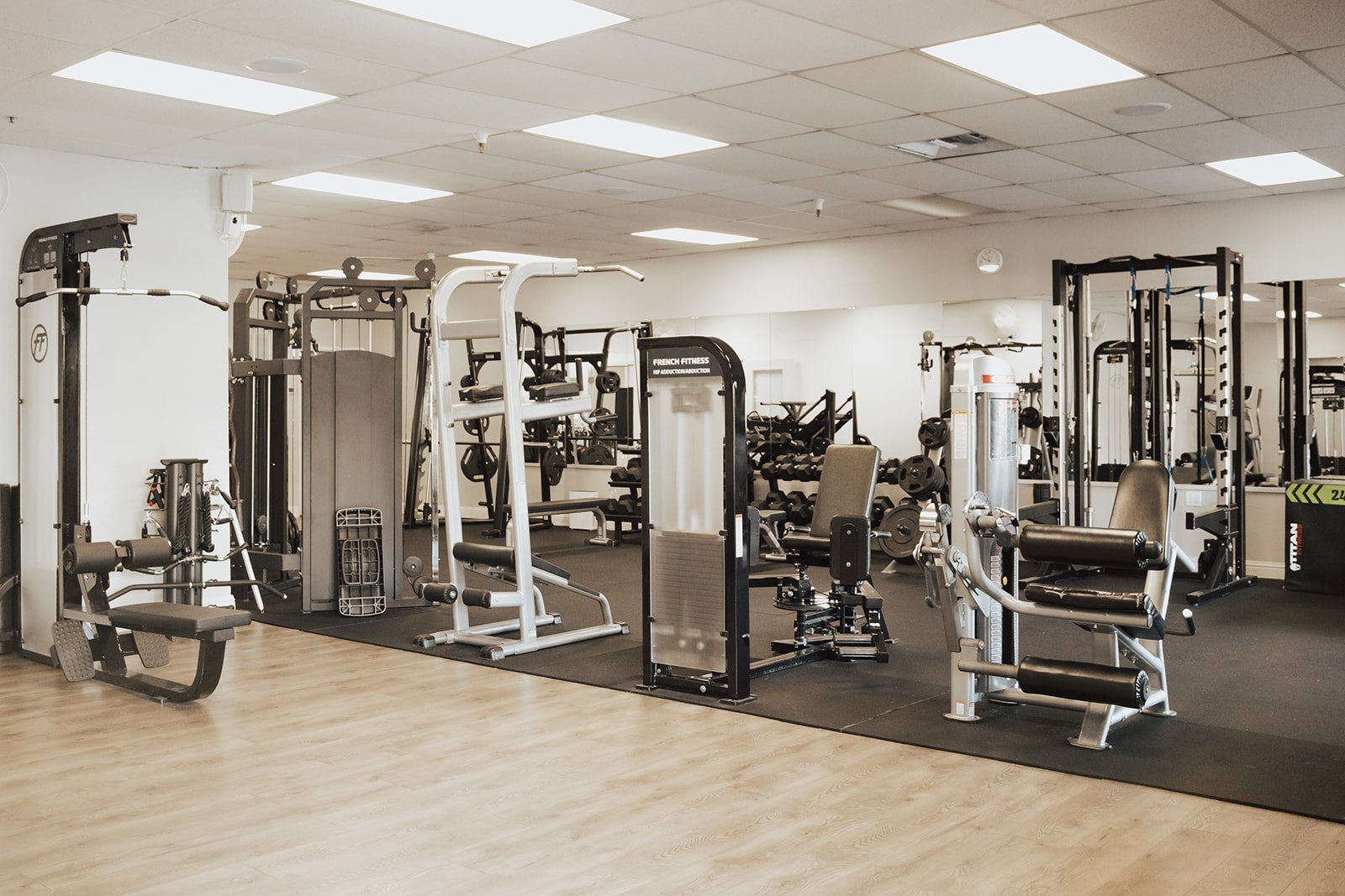 A gym with various strength training equipment including weight machines, free weights, and cardio equipment, with a wood floor and a drop ceiling.