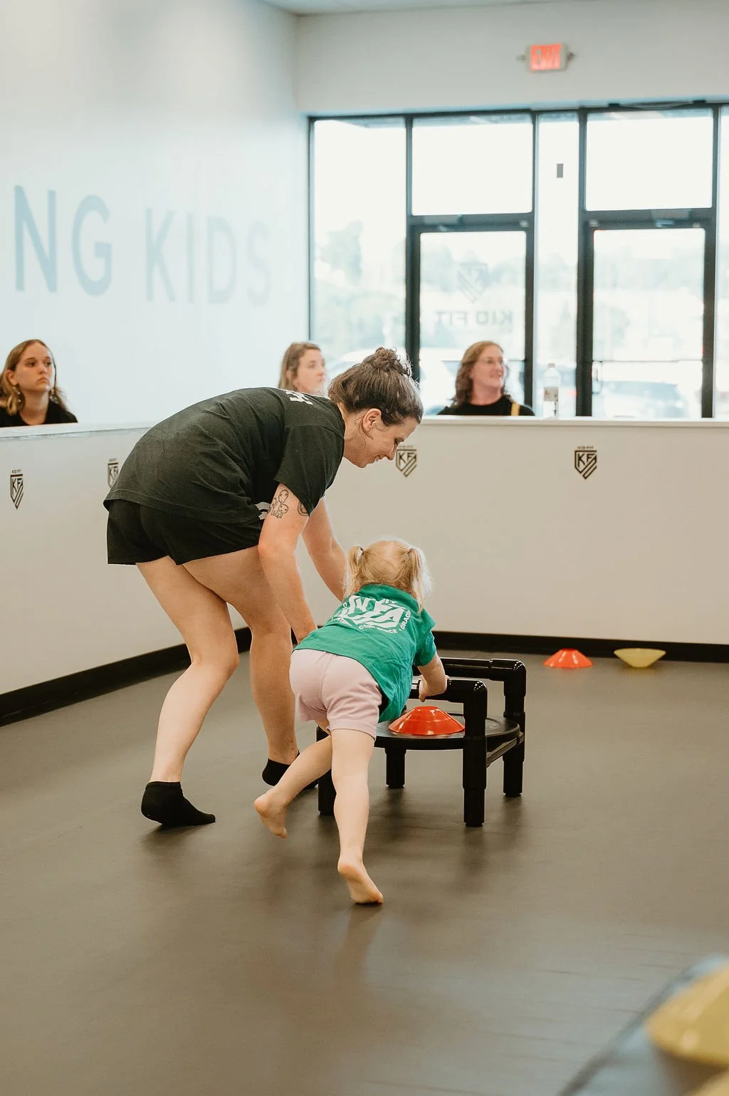 A woman helping a young girl in a green shirt and pink shorts push a sled with orange cones on the floor, with several women sitting at a counter in the background.