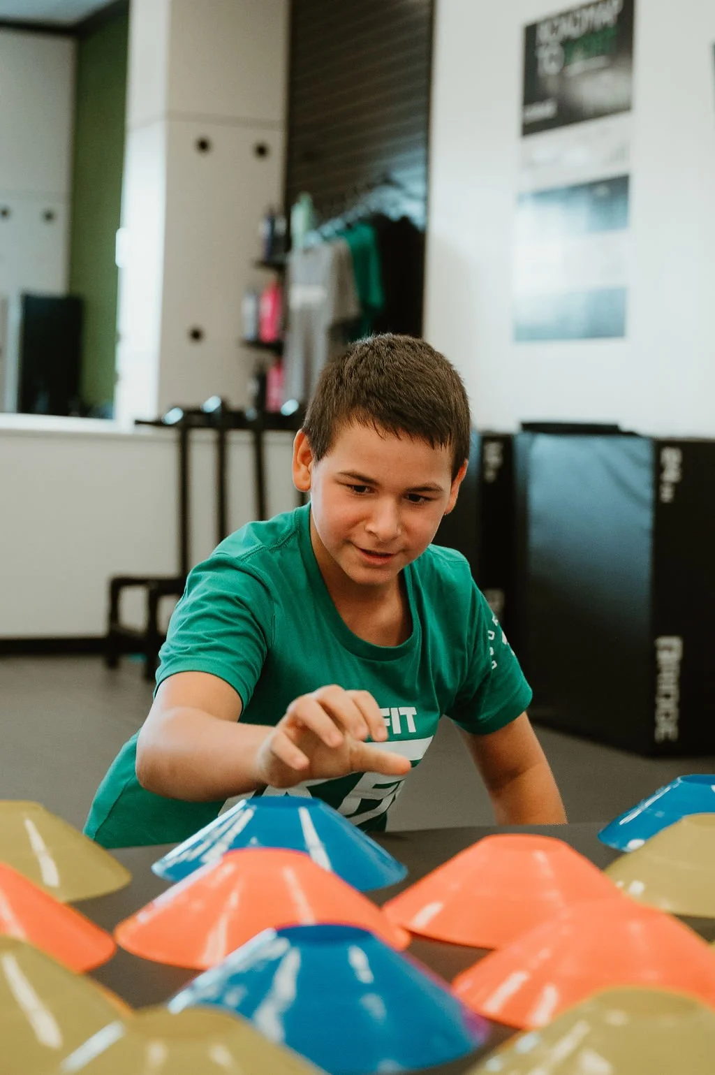A boy in a green Kid Fit shirt playing with colorful cone-shaped objects indoors.