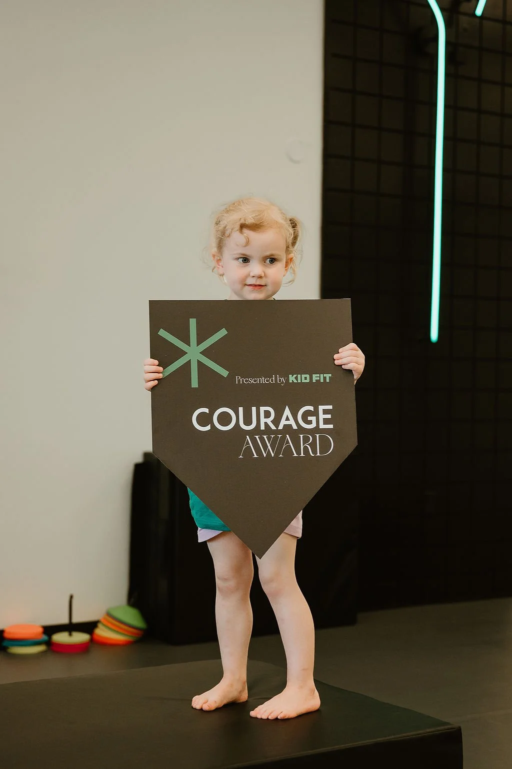 A young girl with curly blonde hair holding a black award sign that says "Courage Award" presented by Kid Fit, standing barefoot on a black platform, indoors with a gray and black wall background.