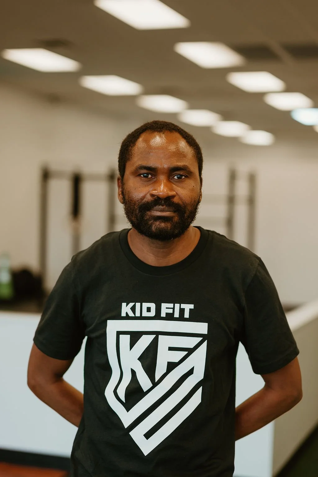 A man with dark hair and a beard wearing a black "Kid Fit" t-shirt standing indoors with a neutral expression.