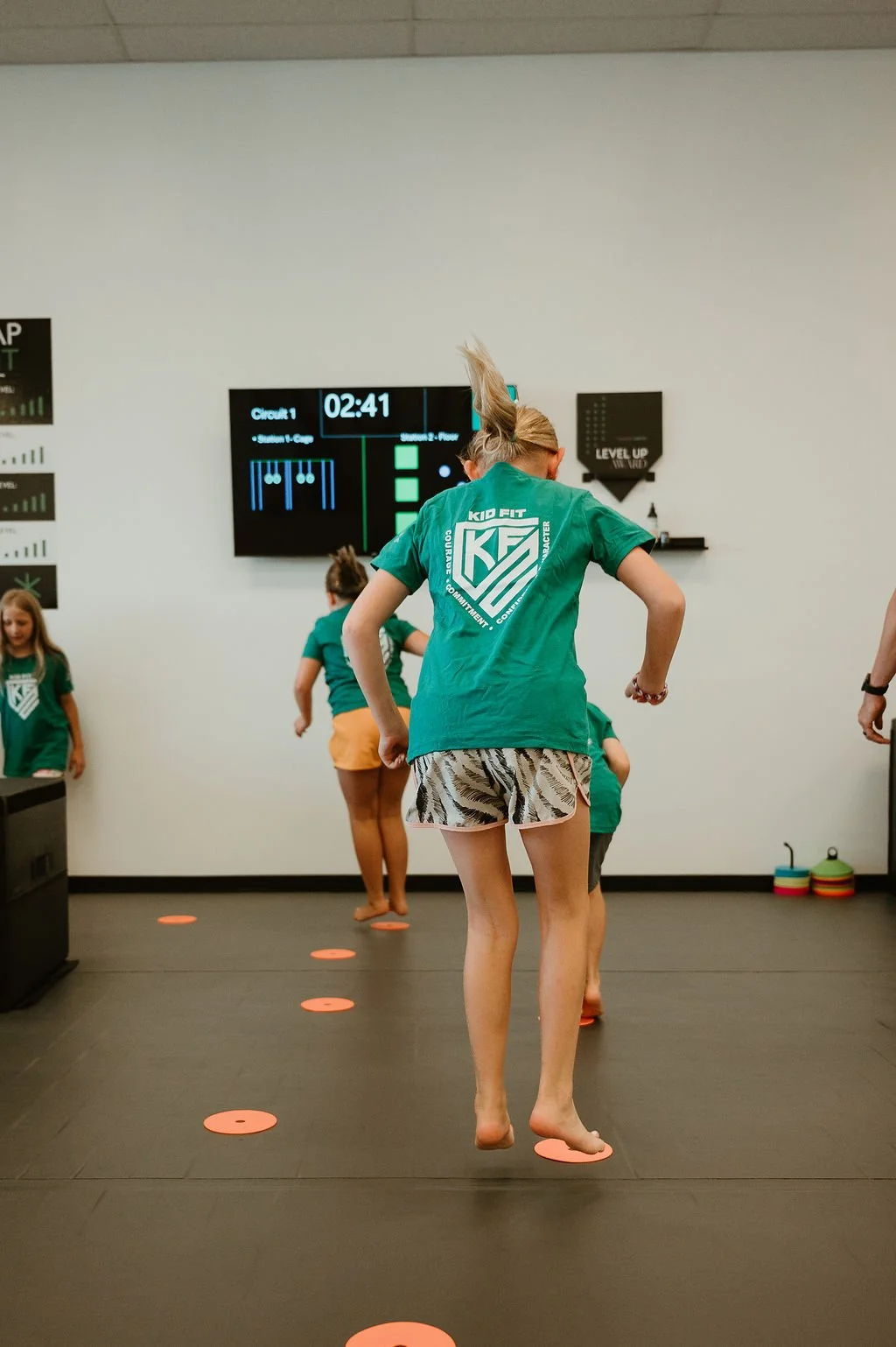 Children in a fitness class stepping on orange discs arranged in a line on the black floor, with a white wall and a digital timer on the wall behind them.