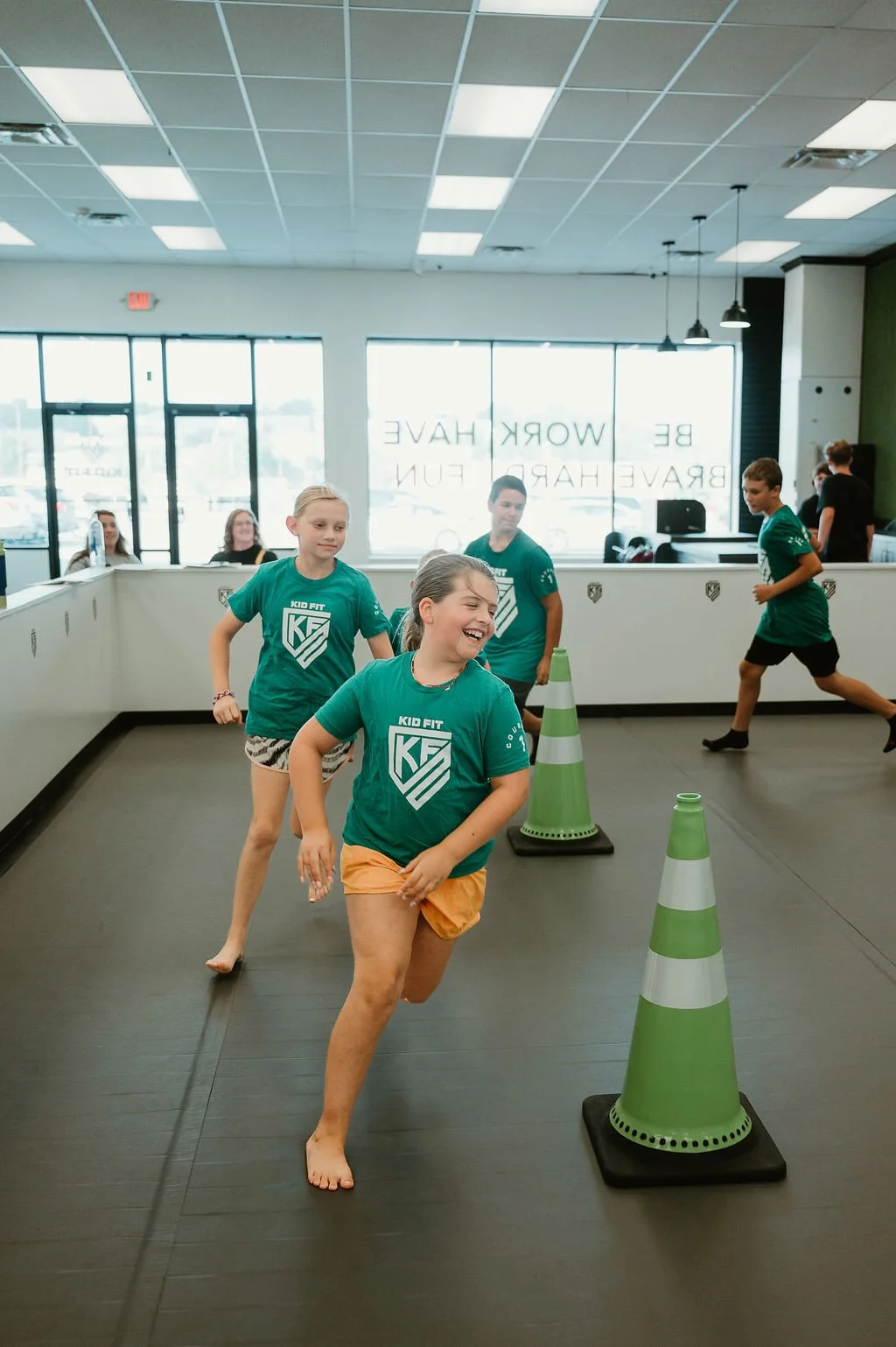 Children participating in a Kid Fit activity indoors, running barefoot around green and white traffic cones.