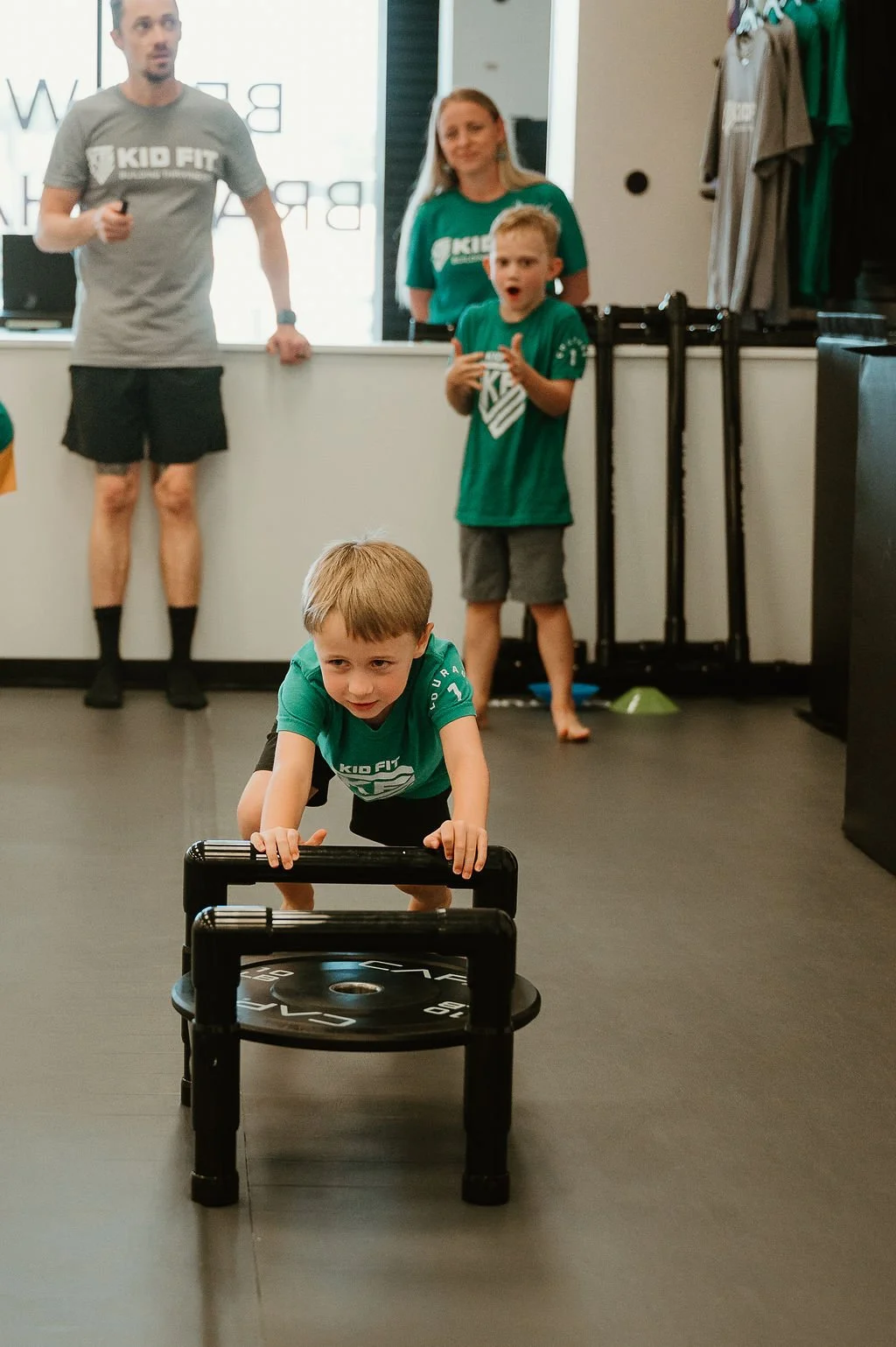 A young boy in a green Kid Fit t-shirt pushes a weight sled across a gym floor. In the background, another boy, a woman, and a man watch, all wearing matching Kid Fit t-shirts.