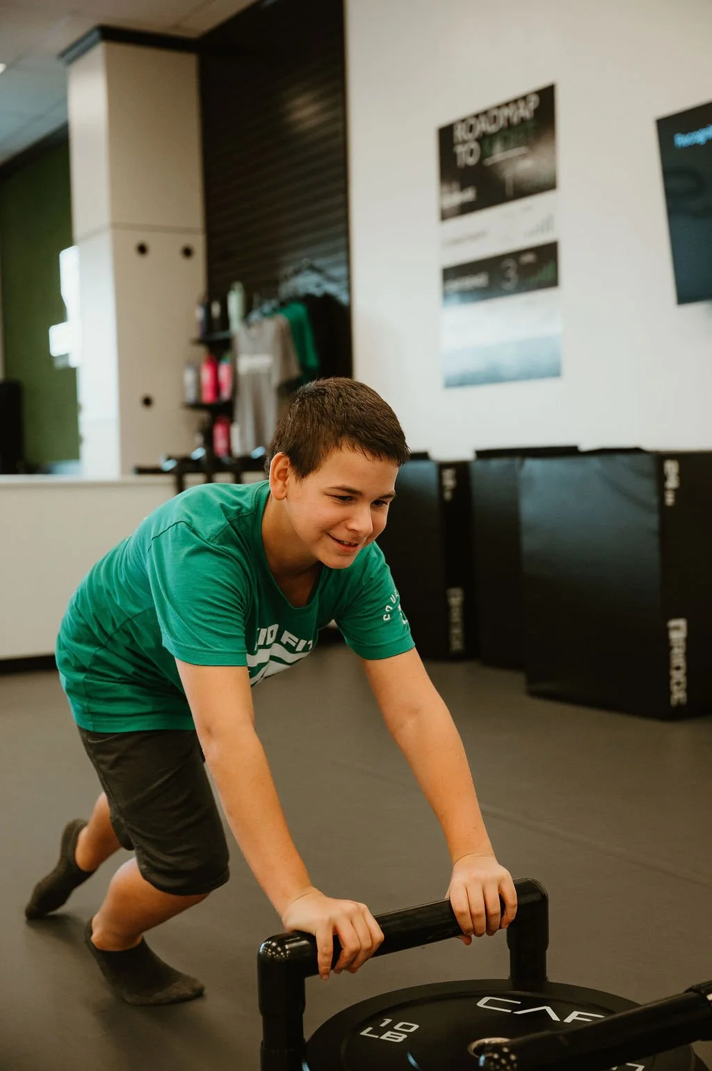 A young boy in a green t-shirt and black shorts smiling as he pushes a weighted sled.