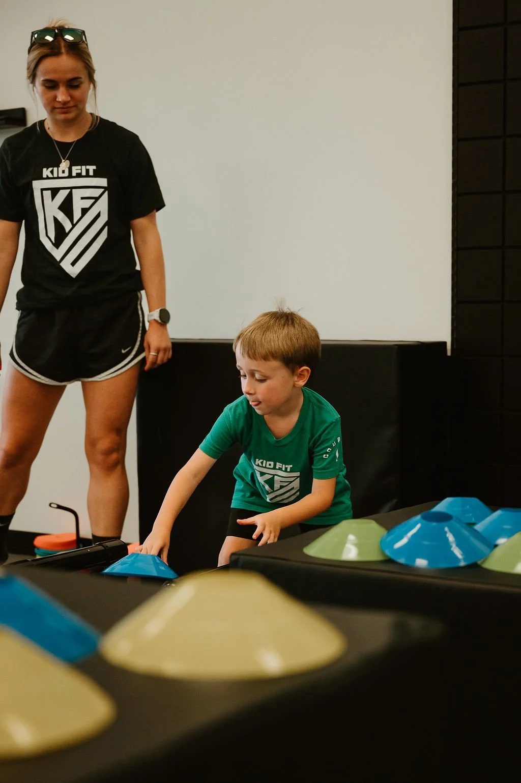 A young boy and a woman at a Kid Fit gym, the boy is reaching for colorful fitness cones on a table, the woman stands nearby watching.