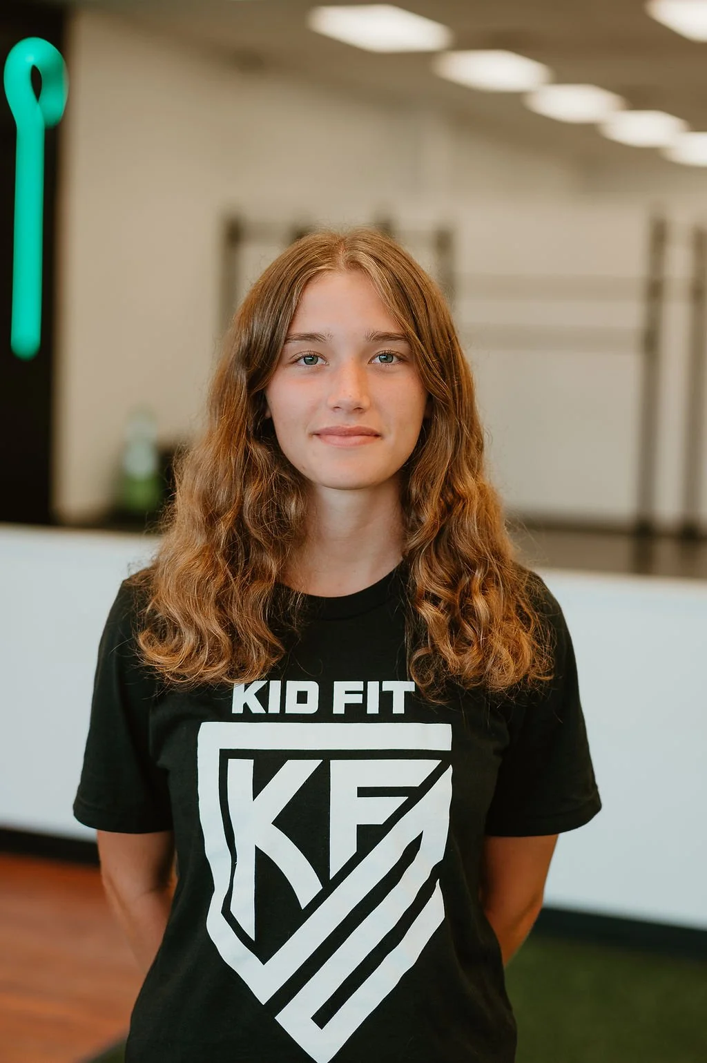 A young girl with long, curly, light brown hair and blue eyes stands indoors with a neutral expression. She is wearing a black T-shirt with white text and a logo that reads "KID FIT." In the background, there are blurred gym equipment and ceiling lights.