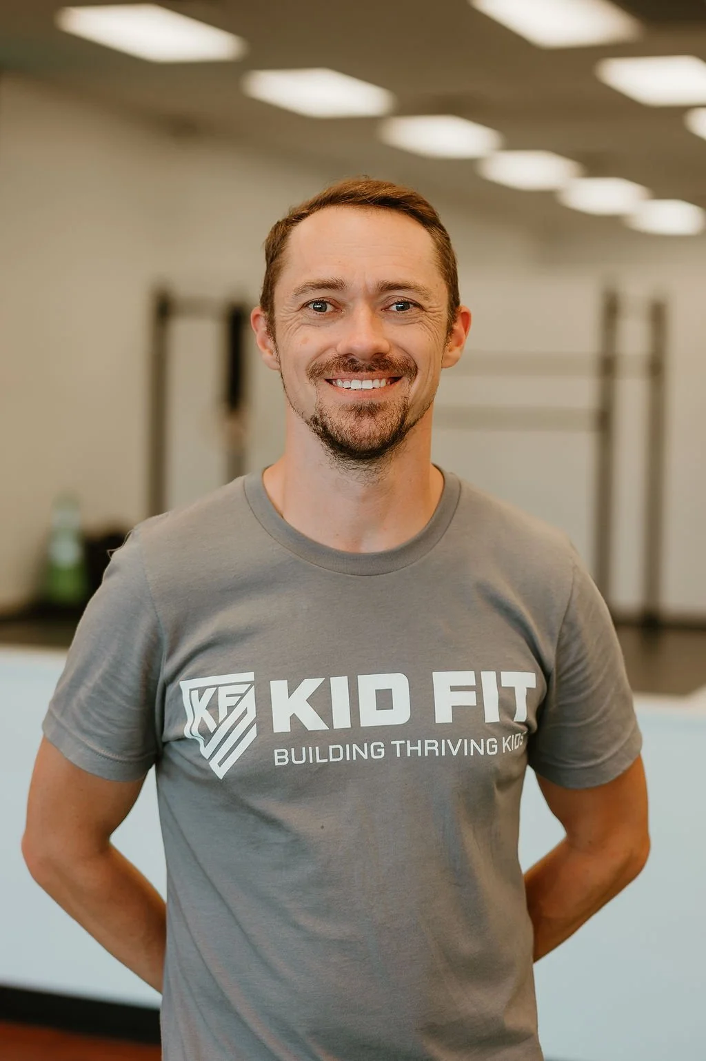 A smiling man wearing a gray T-shirt with the logo 'Kid Fit' and the slogan 'Building Thriving Kids' standing in a gym.