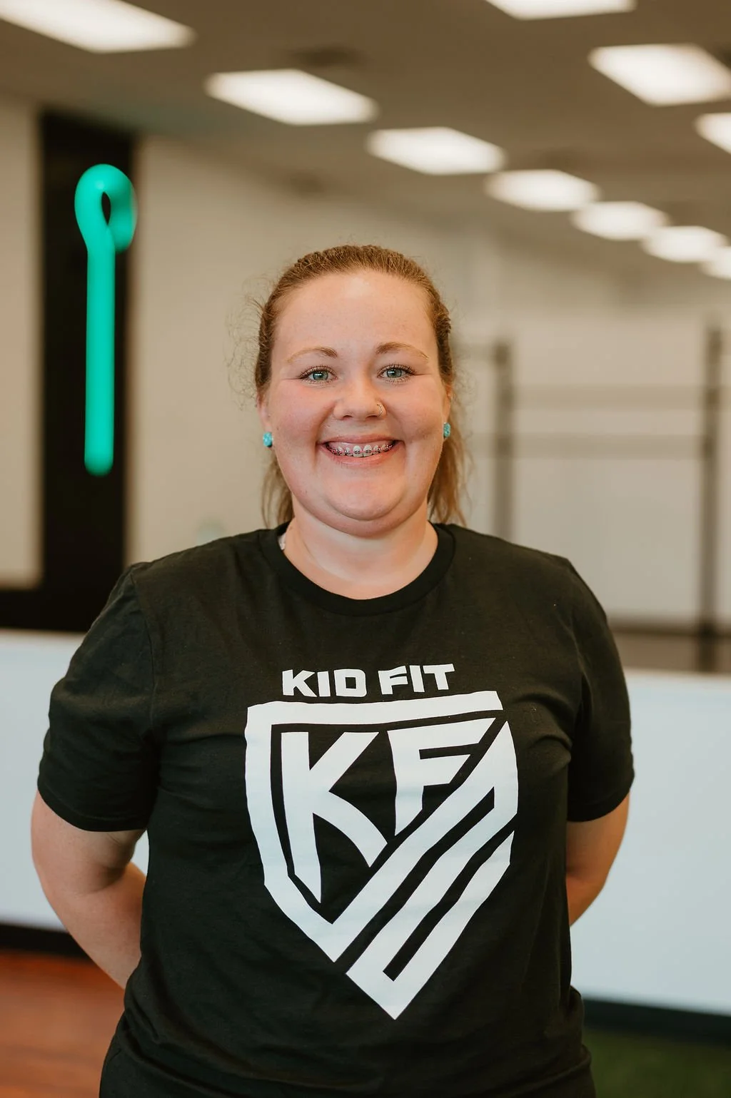 Smiling woman with red hair in a ponytail, wearing a black T-shirt with a white logo that says 'Kid Fit' and blue earrings, standing in an indoor gym or fitness center.