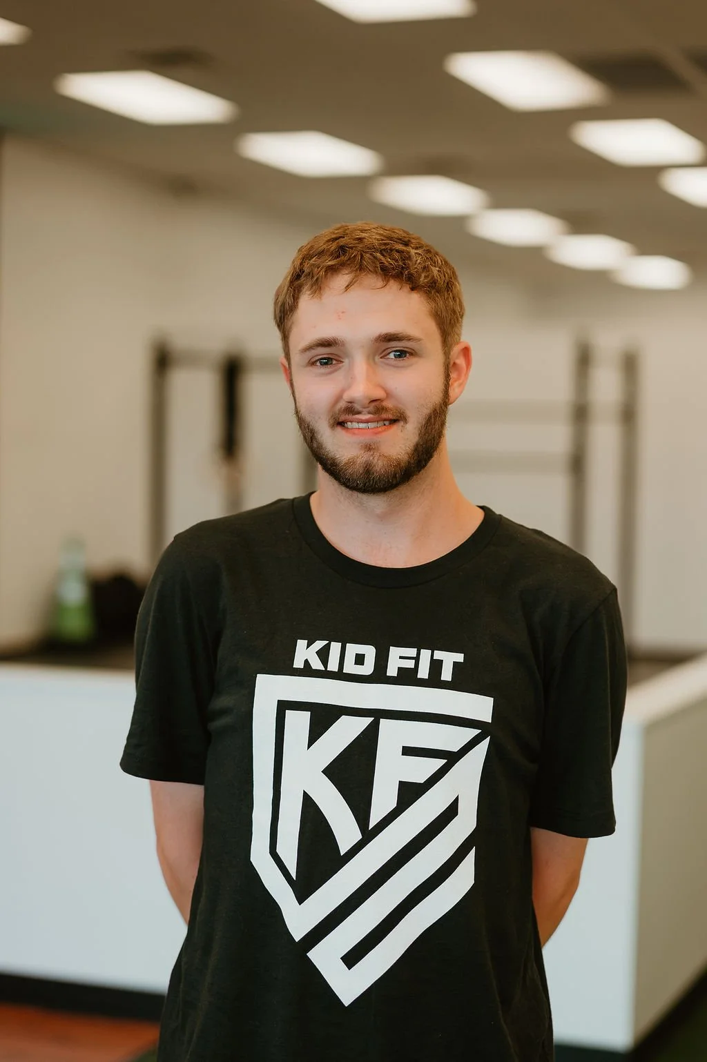 Smiling young man with brown hair and a beard, wearing a black T-shirt with a white logo that says 'Kid Fit', standing in an indoor gym.