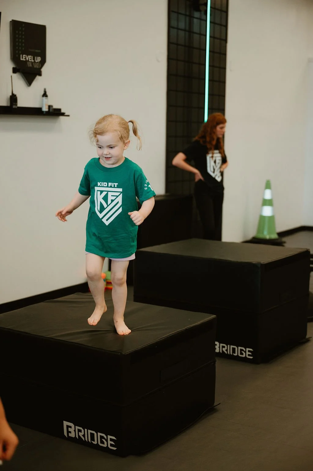 A young girl with blonde hair, wearing a green Kid Fit T-shirt, balances on a black foam step at an indoor fitness facility. She is barefoot and appears happy. An adult woman with curly red hair, also in a Kid Fit T-shirt, stands in the background.