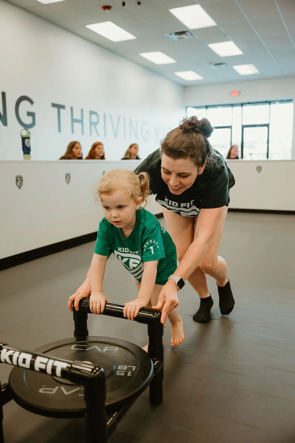 A woman and a young girl with blonde hair and a green KidFit t-shirt are exercising at a gym, pushing a small weight sled together. The woman is guiding the girl, who is barefoot, on a dark gym floor, with a white wall and large windows in the backgr