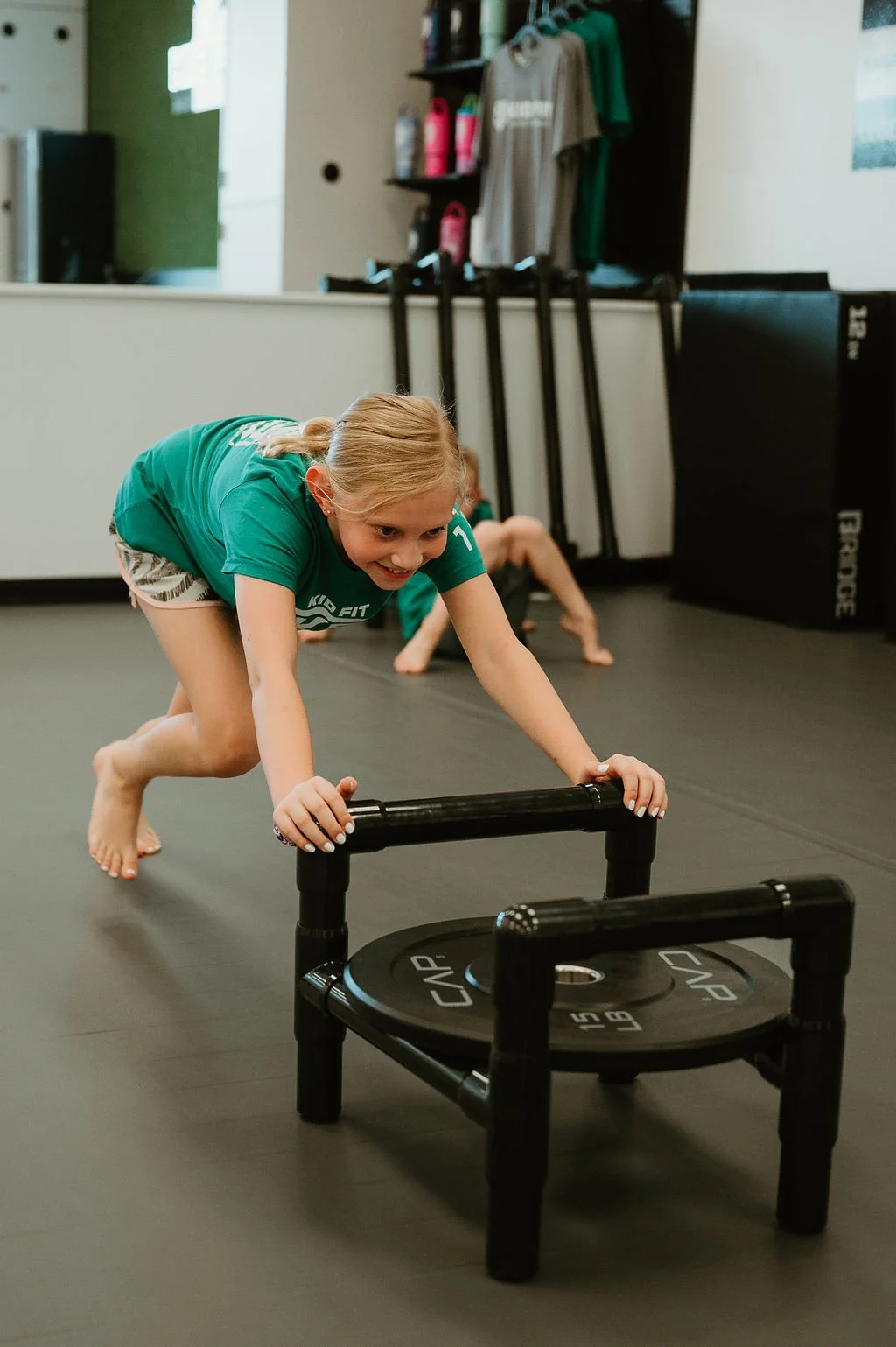 Children pushing sleds at a gym, with one girl in the foreground smiling, wearing a green Kid Fit T-shirt and shorts.