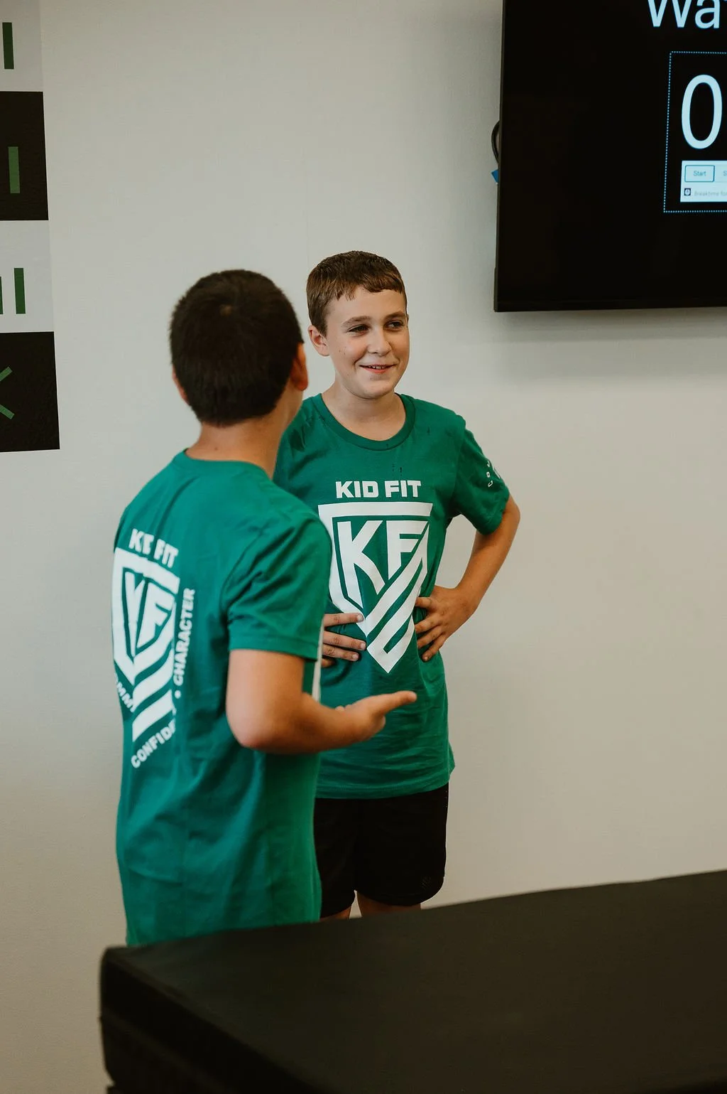 Two boys wearing green t-shirts with 'Kid Fit' and a shield logo are talking and smiling indoors, with a white wall and a digital screen in the background.