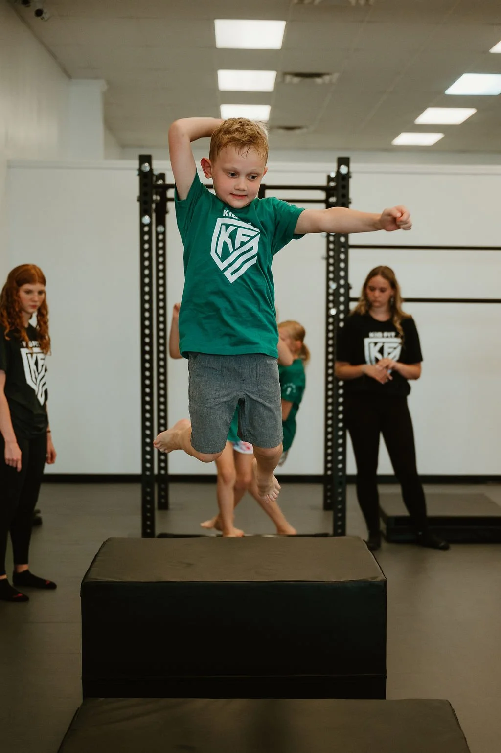 A young boy jumping onto gym mats in an indoor gym, with other children and a woman observing, all wearing shirts with a Kid Fit logo.