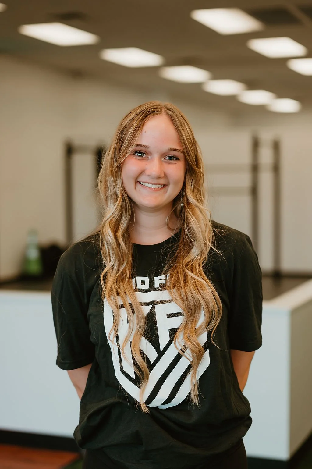 Young woman with long wavy blonde hair smiling and standing indoors in a black T-shirt with a white graphic design.