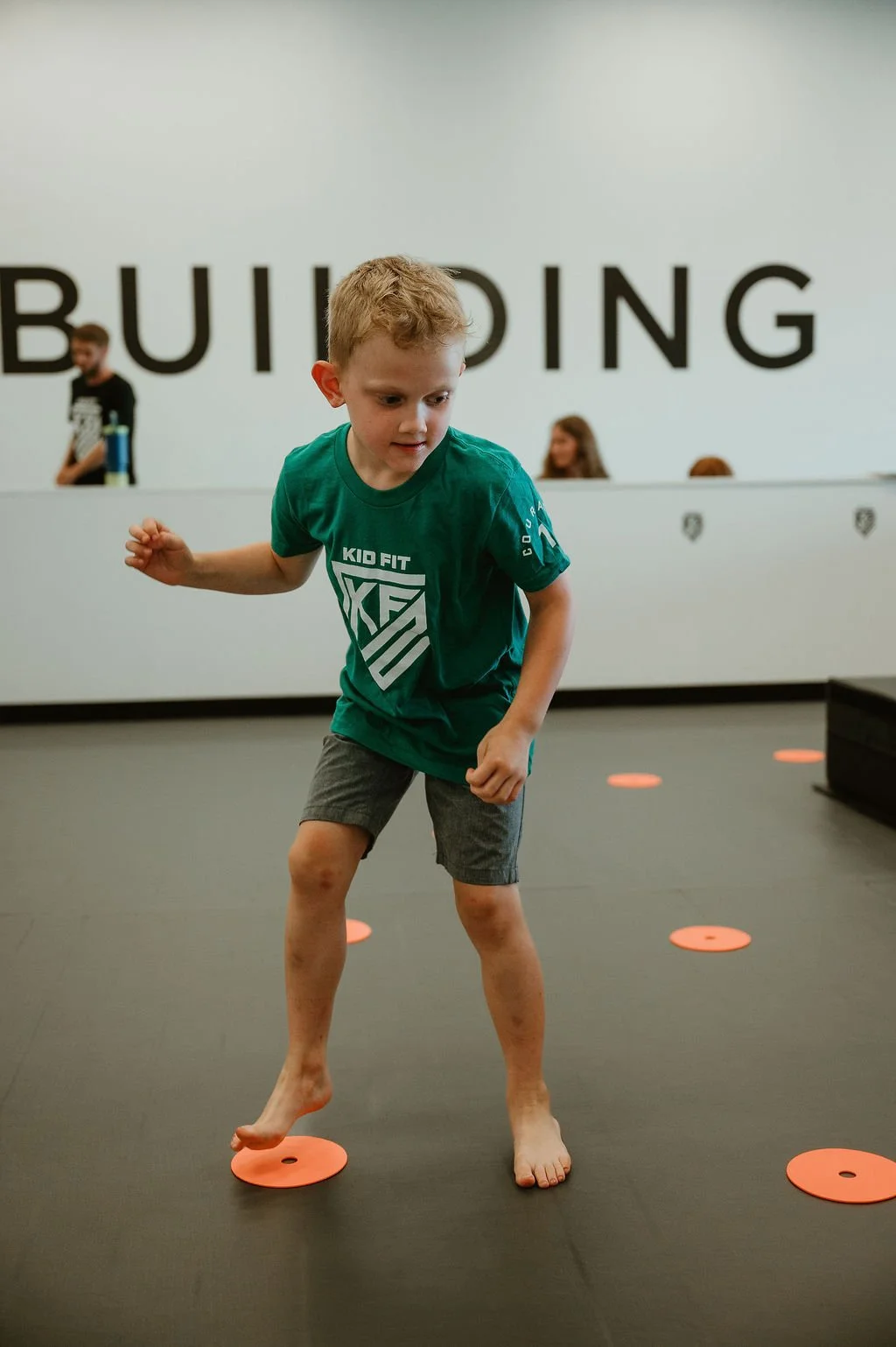 A young boy with blonde hair in a green T-shirt and gray shorts is balancing barefoot on orange circular discs on a black floor in a gym or indoor play area. The background features the large text 'BUILDING' on a white wall.