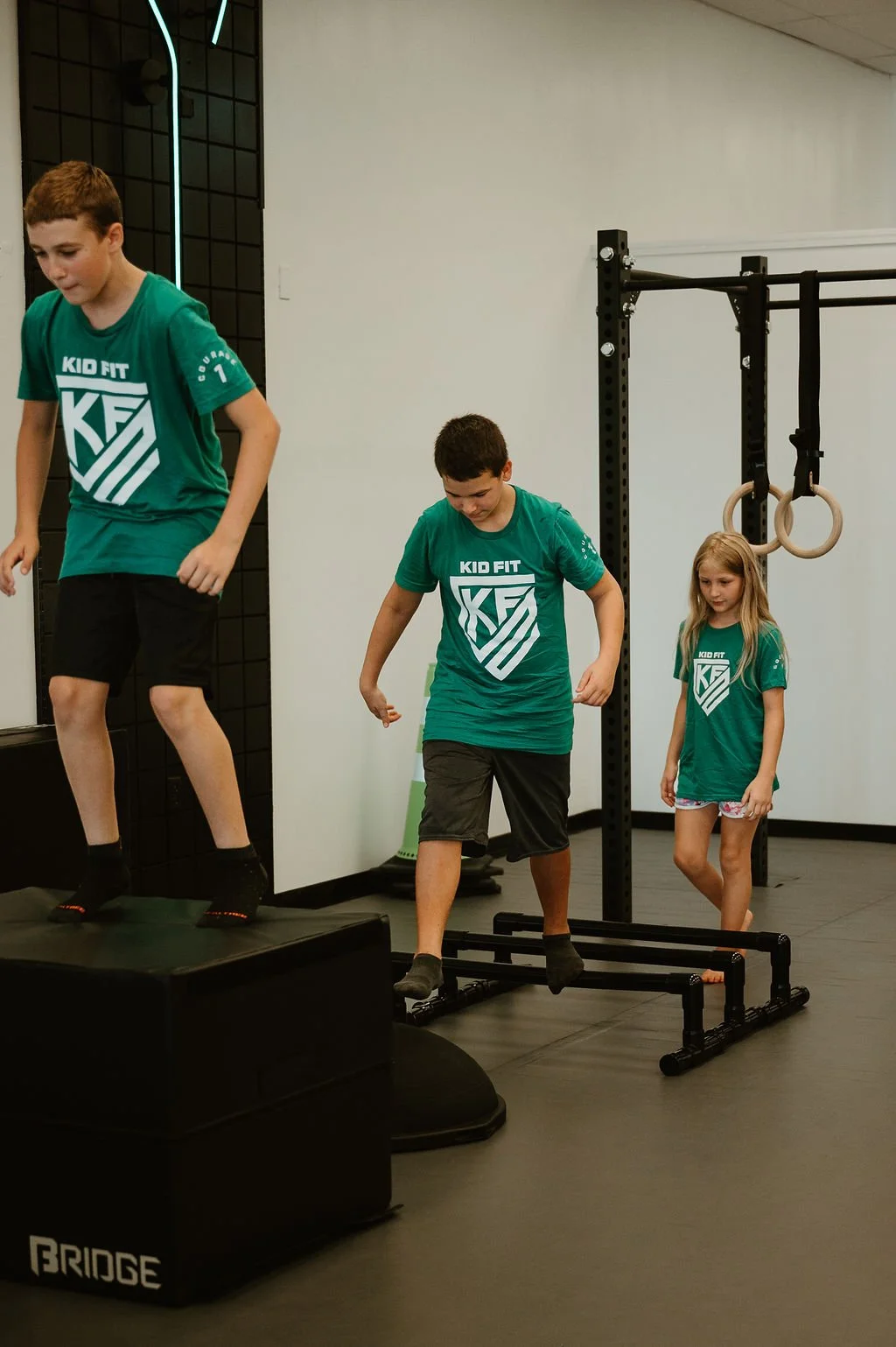 Three children in green 'Kid Fit' shirts participating in an indoor fitness activity, with one child jumping onto a raised platform, another stepping onto a jump box, and a young girl standing nearby, in a gym with gymnastic rings and a black metal s