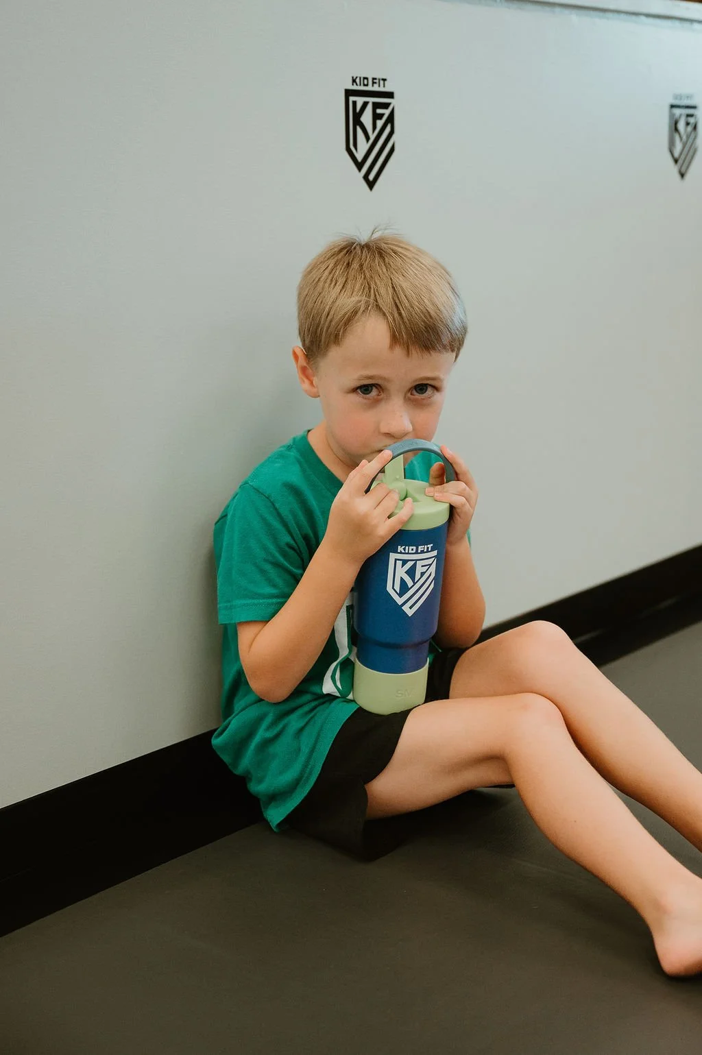 A young boy with short blonde hair sitting on the floor against a wall, holding a blue and green Kid Fit water bottle, looking at the camera.